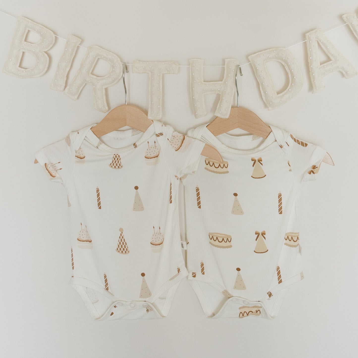 Two white "Short Sleeve Bamboo Snapsuits | Birthday Boy" by forever french baby, crafted from breathable bamboo fabric and featuring birthday-themed prints, hang on wooden hangers. Above them is a cream cloth "BIRTHDAY" banner, all set against a plain white background.