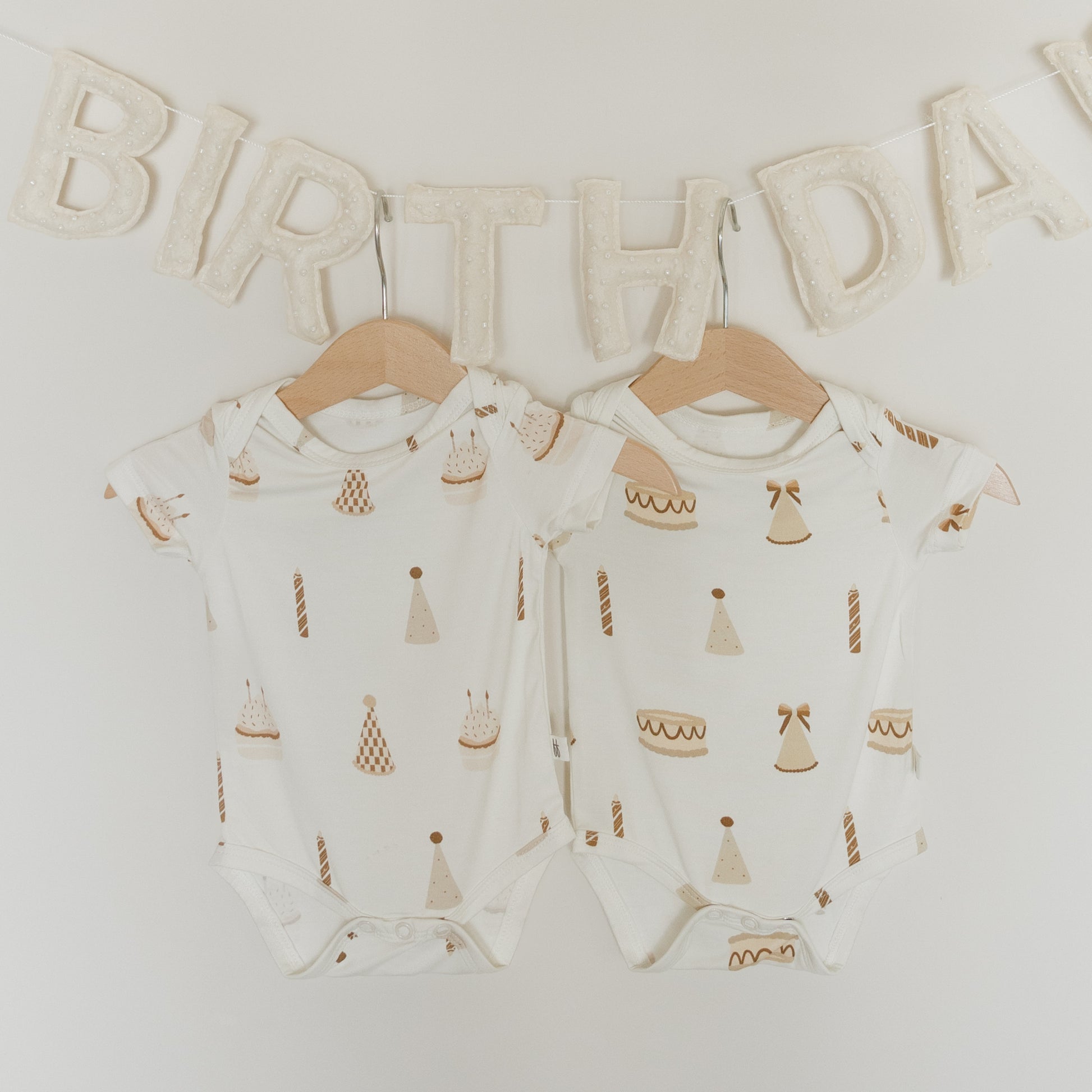 Two white "Short Sleeve Bamboo Snapsuits | Birthday Boy" by forever french baby, crafted from breathable bamboo fabric and featuring birthday-themed prints, hang on wooden hangers. Above them is a cream cloth "BIRTHDAY" banner, all set against a plain white background.