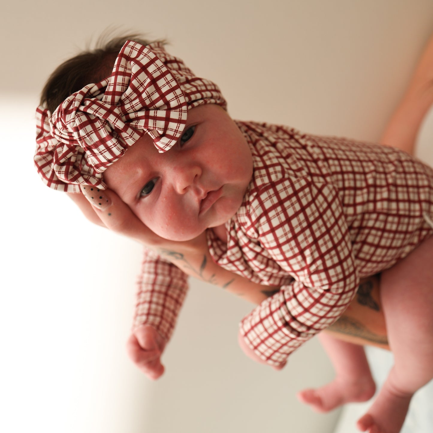 A baby wears the forever french baby Long Sleeve Bamboo Snapsuit in Window Pane, complete with a matching headband and bow, held by an adult with tattooed arms, gazing at the camera against a plain background.
