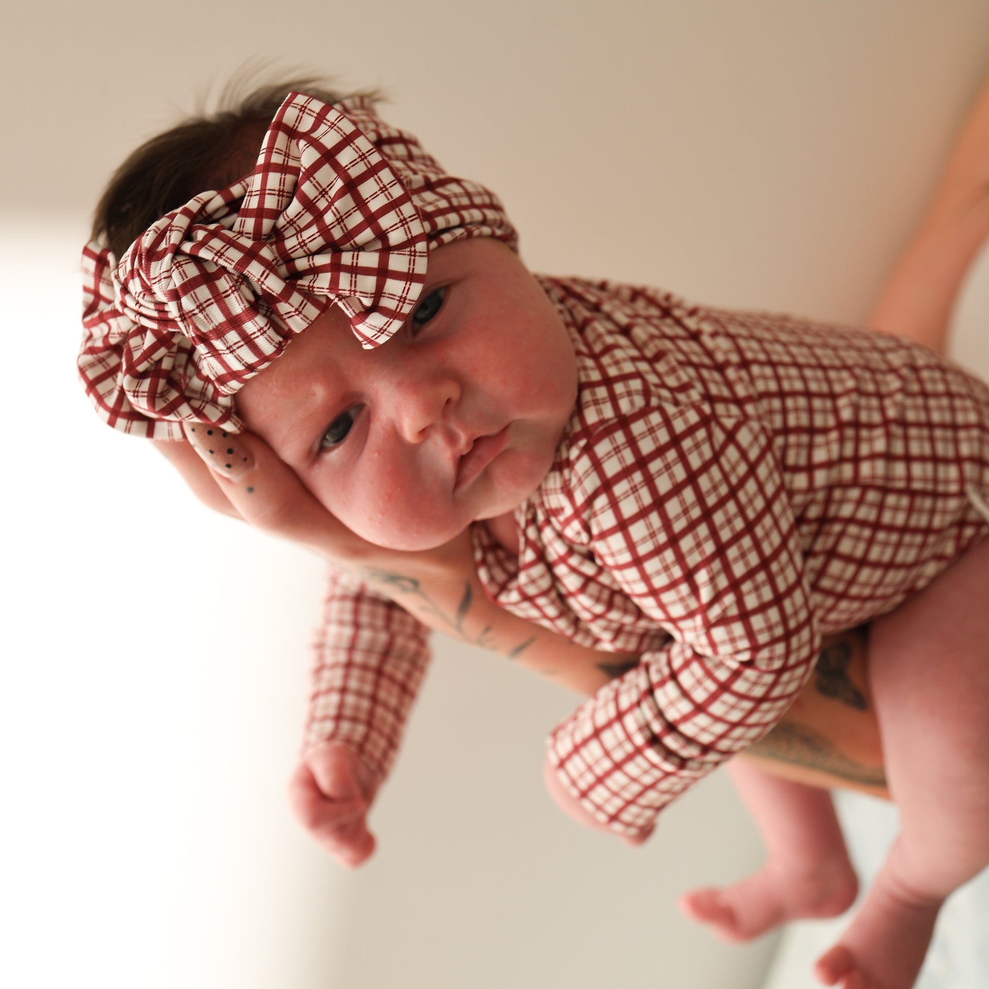 A baby wears the forever french baby Long Sleeve Bamboo Snapsuit in Window Pane, complete with a matching headband and bow, held by an adult with tattooed arms, gazing at the camera against a plain background.