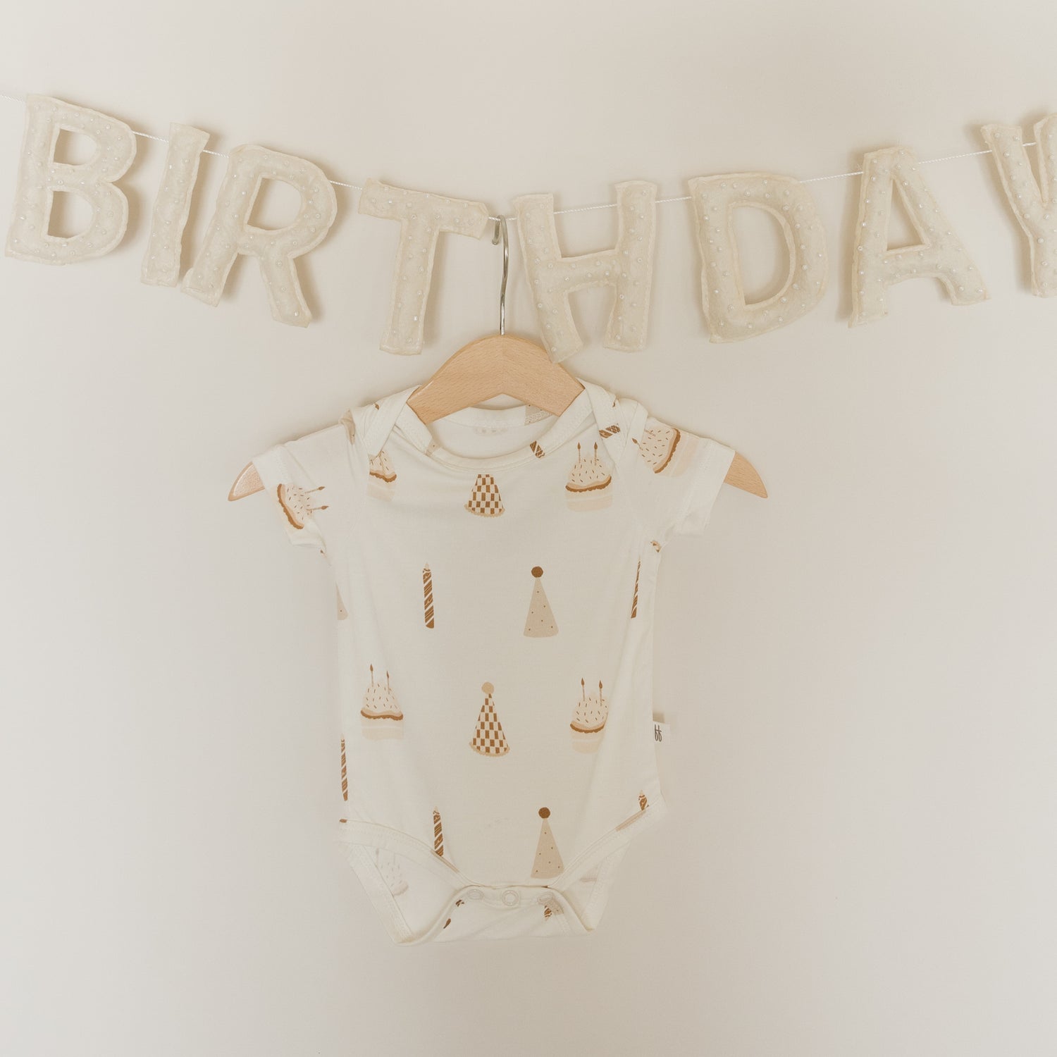 A forever french baby Short Sleeve Bamboo Snapsuit | Birthday Boy in white, featuring playful birthday cake and candle prints, hangs on a wooden hanger beneath a soft, textured "BIRTHDAY" banner. This hypoallergenic snapsuit combines adorable style with the breathable comfort of bamboo fabric—perfect for your little one’s special celebration.
