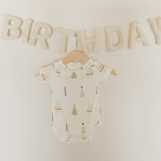 A forever french baby Short Sleeve Bamboo Snapsuit | Birthday Boy in white, featuring playful birthday cake and candle prints, hangs on a wooden hanger beneath a soft, textured "BIRTHDAY" banner. This hypoallergenic snapsuit combines adorable style with the breathable comfort of bamboo fabric—perfect for your little one’s special celebration.