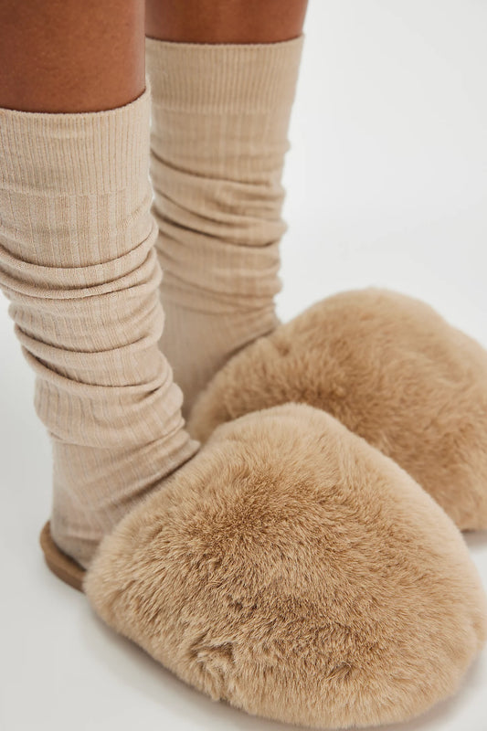 Someone wears thick beige ribbed socks with oversized, fluffy tan Free People Snowball Slippers while standing on a white surface.