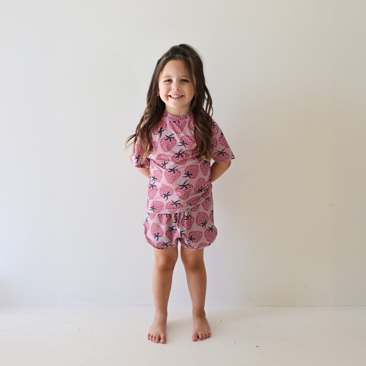 A young girl with long brown hair smiles barefoot by a white wall, wearing the forever french baby Girl's Bamboo Short Set | Matilda—soft, stylish, hypo-allergenic pink pajamas with a black and pink pineapple pattern.