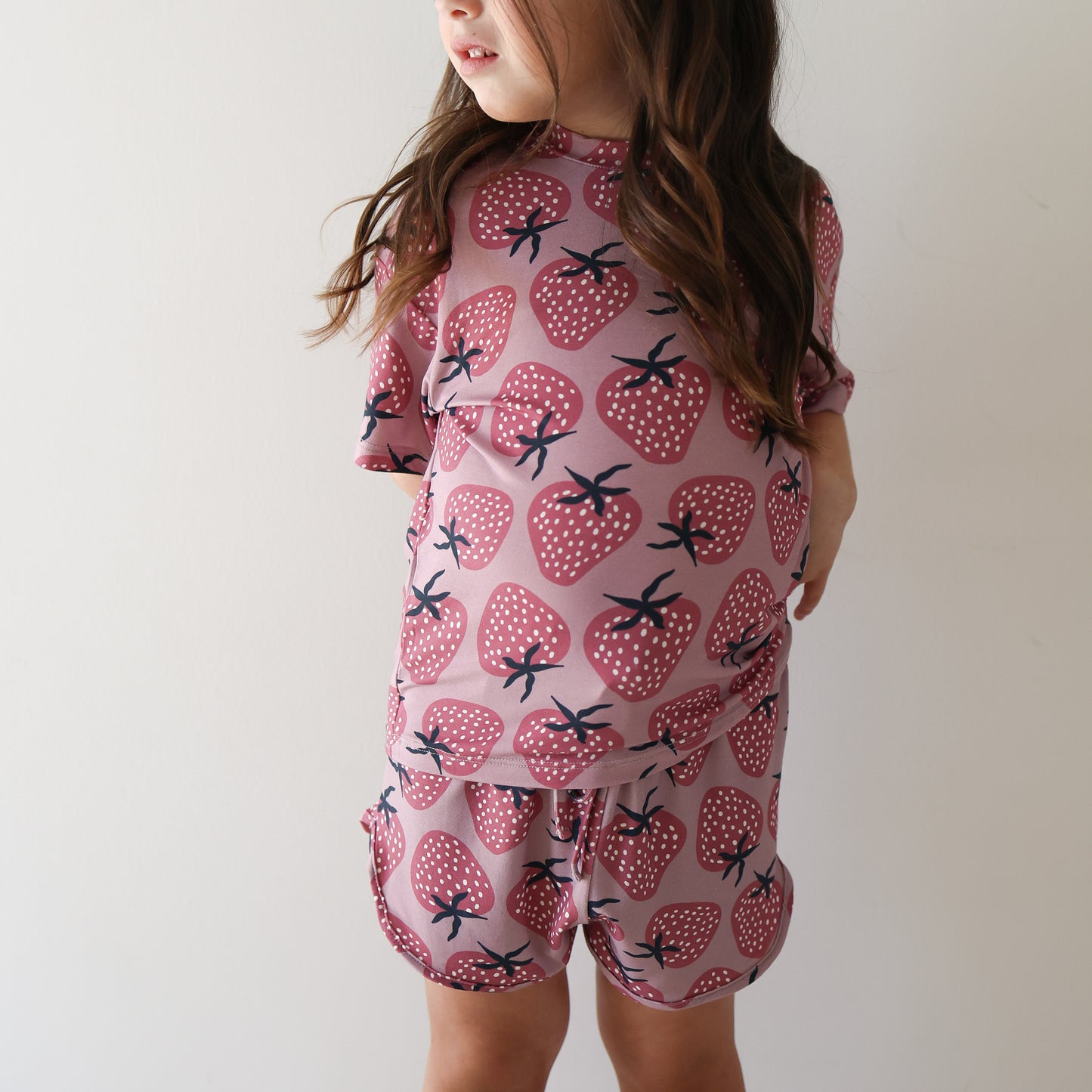 A young girl with long brown hair wears the hypo-allergenic Girl's Bamboo Short Set | Matilda by forever french baby, featuring a pink fabric with red strawberry pattern, standing against a white wall and looking to the side.