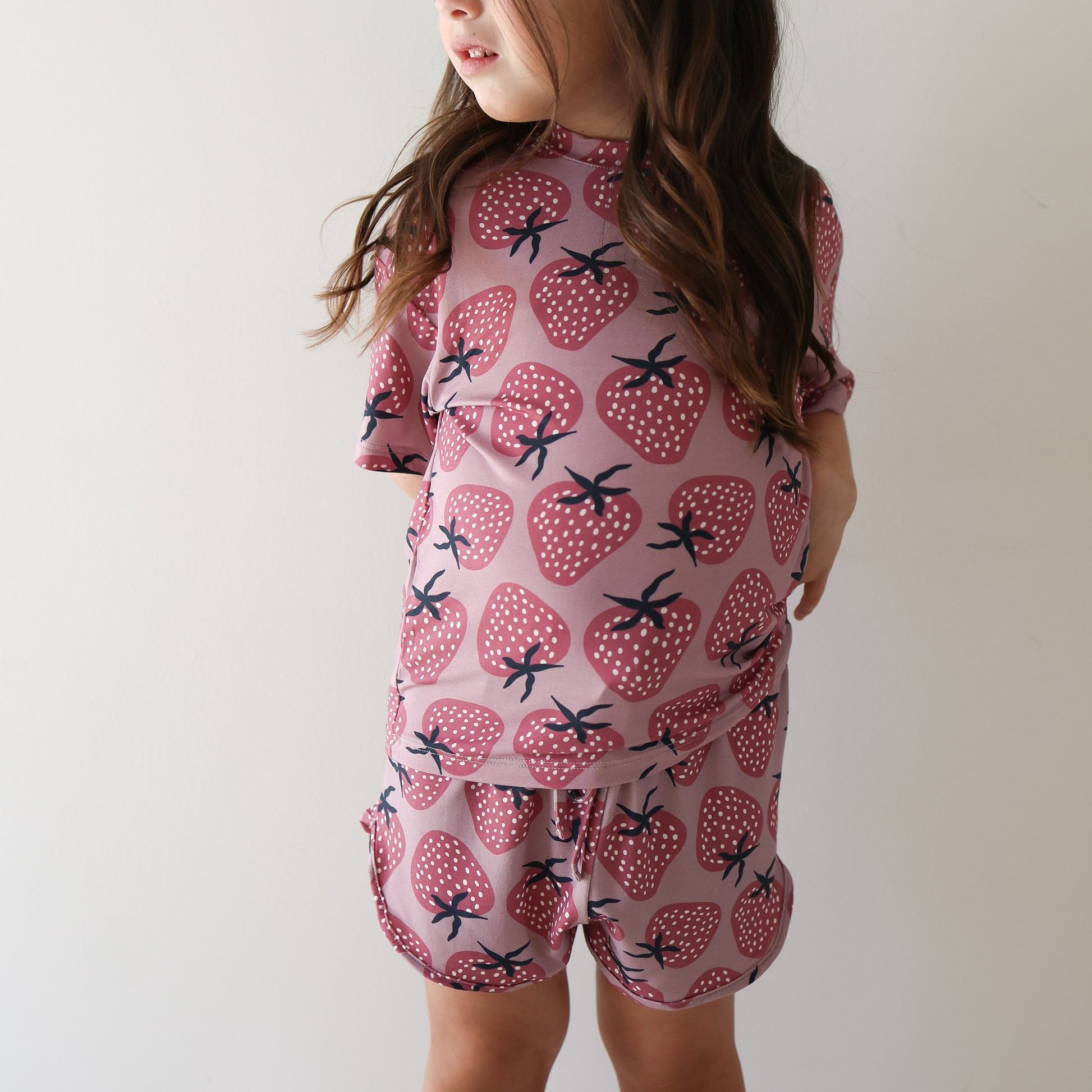 A young girl with long brown hair wears the hypo-allergenic Girl's Bamboo Short Set | Matilda by forever french baby, featuring a pink fabric with red strawberry pattern, standing against a white wall and looking to the side.