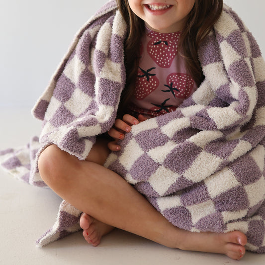 A young girl sits on the floor, smiling and barefoot, wrapped in the forever french baby Plush Blanket | Grape Soda—a soft, purple-and-white checkered microfiber blanket. She wears a shirt with a strawberry pattern.