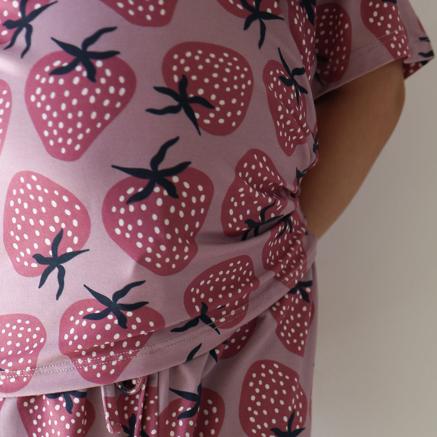 Close-up of a person wearing the Girl's Bamboo Short Set | Matilda by forever french baby, featuring a pink and purple strawberry pattern, showing their torso and arm with hand tucked behind their back.