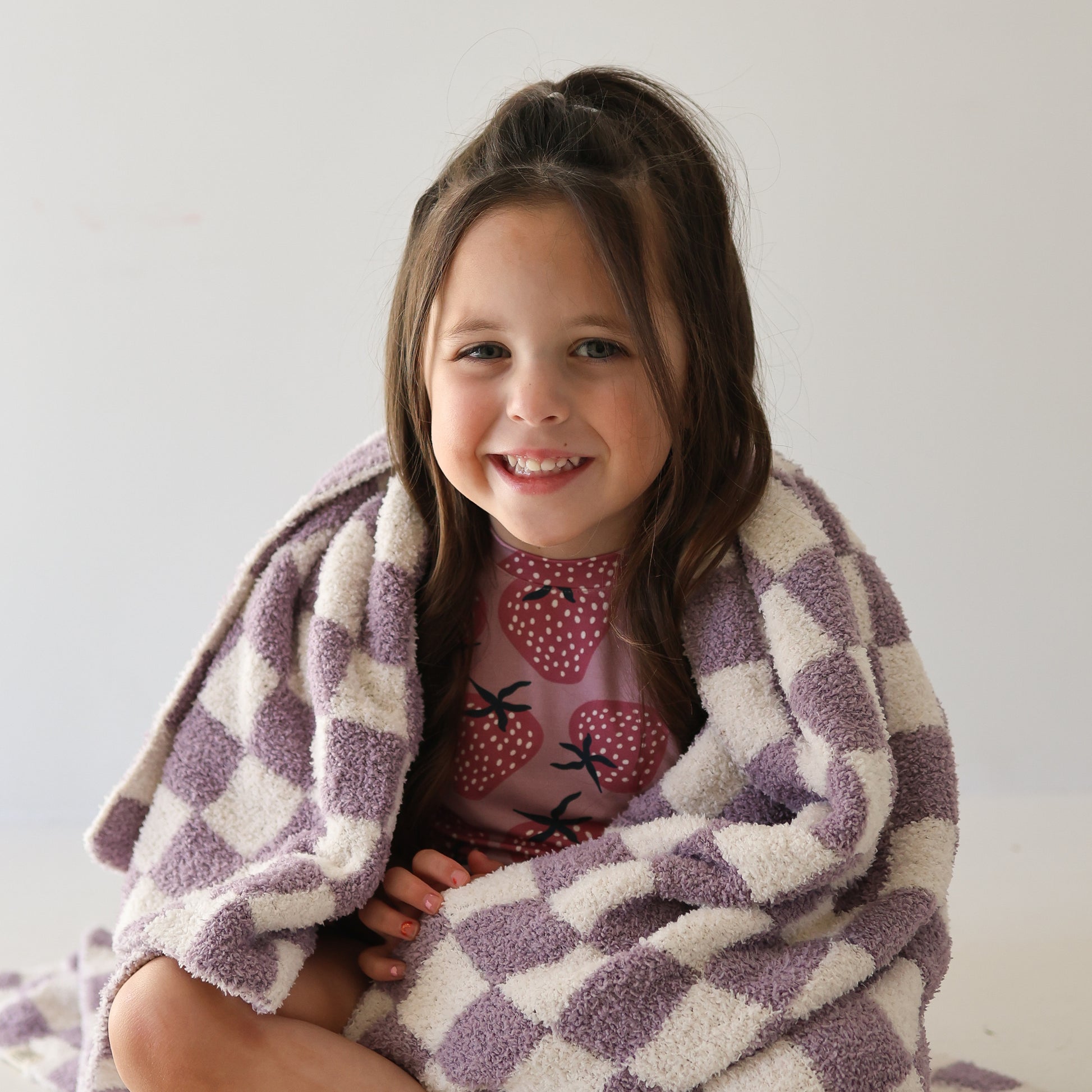 A young girl with long brown hair smiles while wearing the forever french baby Girl’s Bamboo Short Set | Matilda, featuring strawberry prints, wrapped in a purple and white checkered blanket against a plain light background.