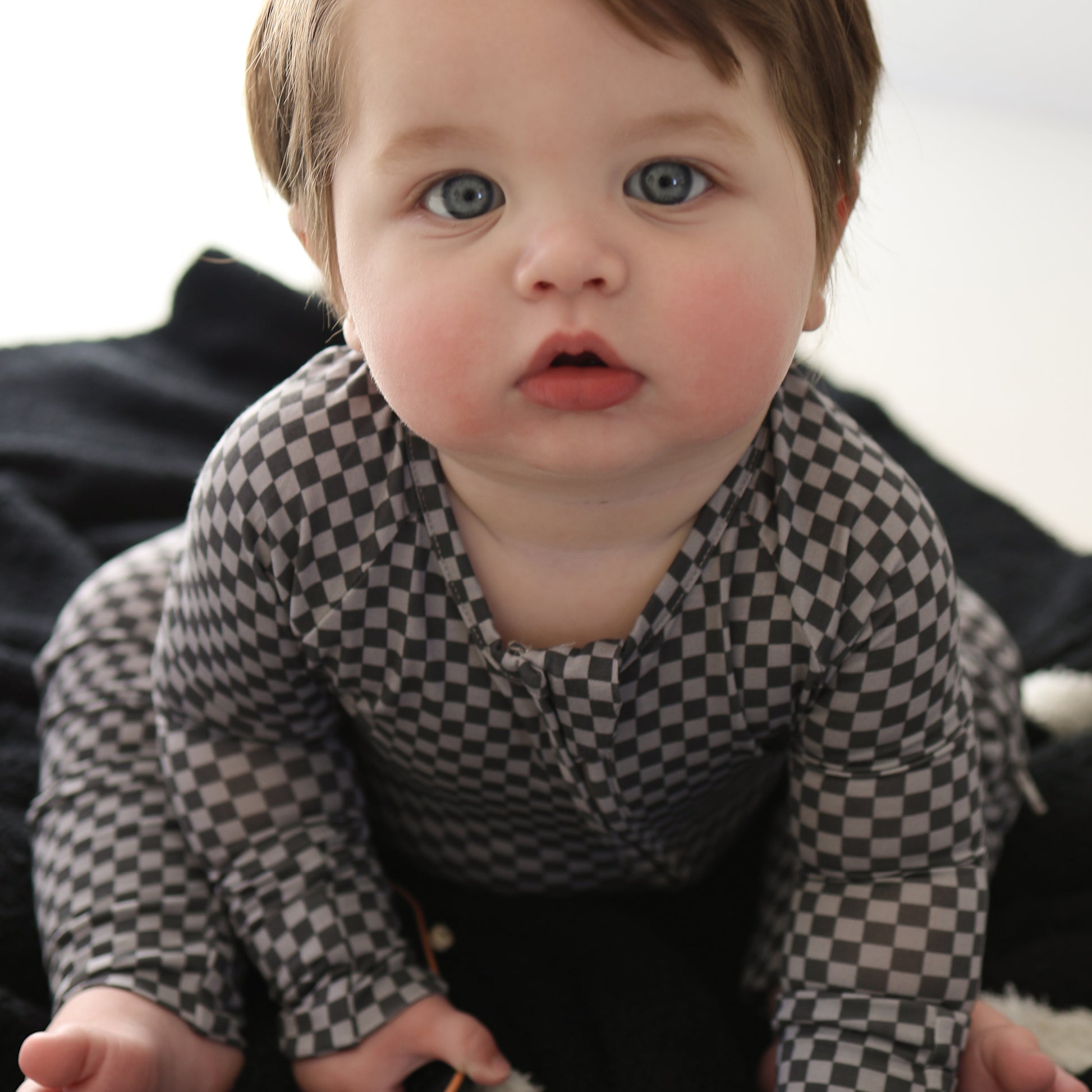 A light-skinned baby with blue eyes and brown hair sits on a black blanket, wearing forever french baby Bamboo Zip Pajamas in the Street Micro Check pattern, looking up with a curious expression.