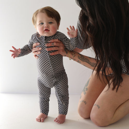A baby wearing forever french baby Bamboo Zip Pajamas in Street Micro Check stands with support from a kneeling adult, who has long dark hair and visible tattoos on their arms and legs.