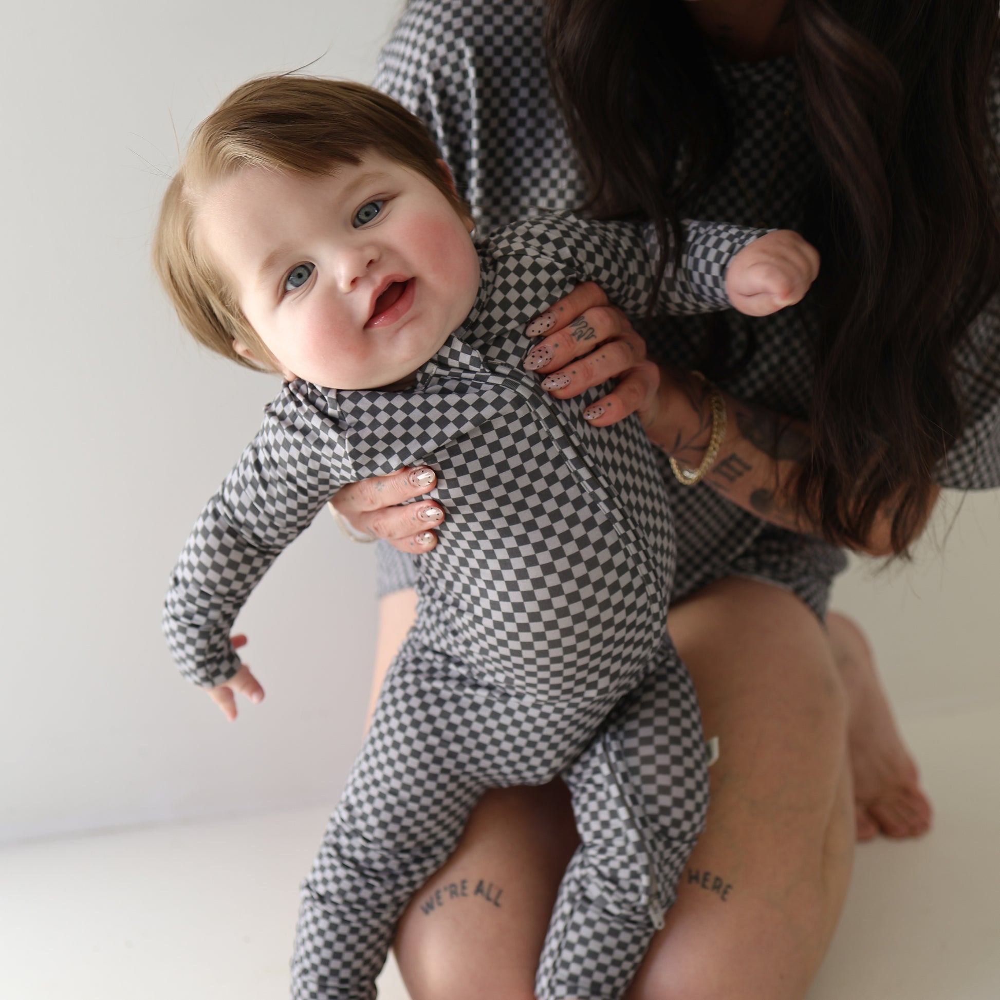 A baby wearing forever french baby's Bamboo Zip Pajamas in Street Micro Check smiles and leans forward while being held by a tattooed adult in matching attire; the adult’s face is partially out of frame.