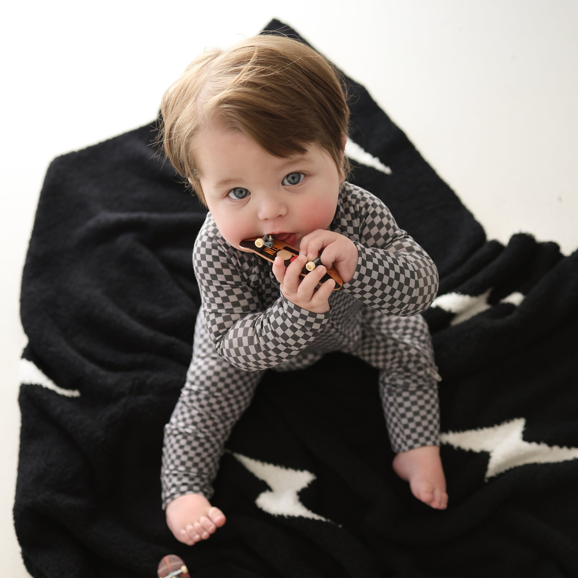 A baby with light brown hair wears forever french baby's Bamboo Zip Pajamas | Street Micro Check, sitting on a black blanket with white stars and chewing a small wooden toy while looking up at the camera.
