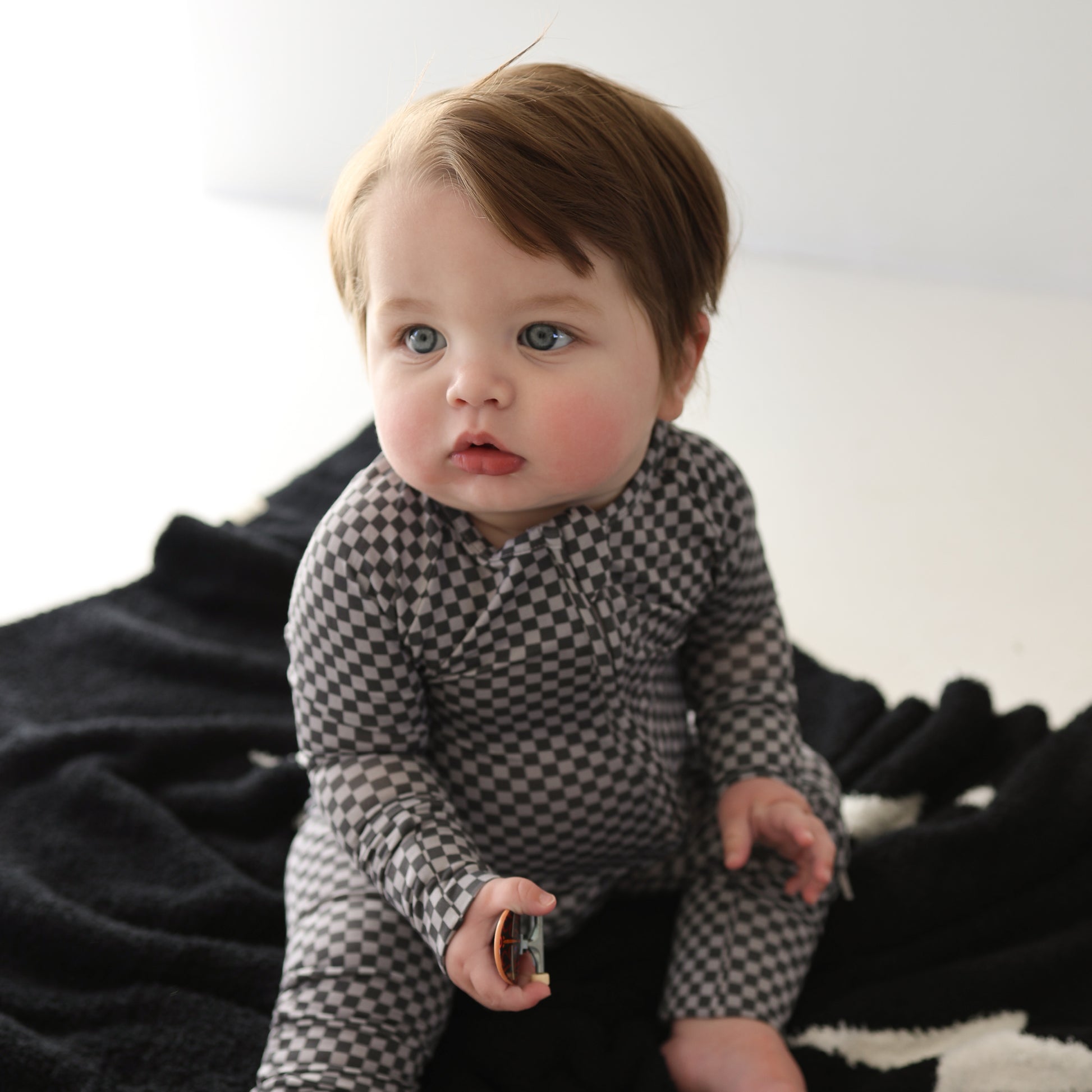A light-skinned baby with blue eyes and brown hair sits on a black blanket, wearing forever french baby's Bamboo Zip Pajamas | Street Micro Check in a gray and black checkered pattern, holding a small round object against a plain, well-lit background.