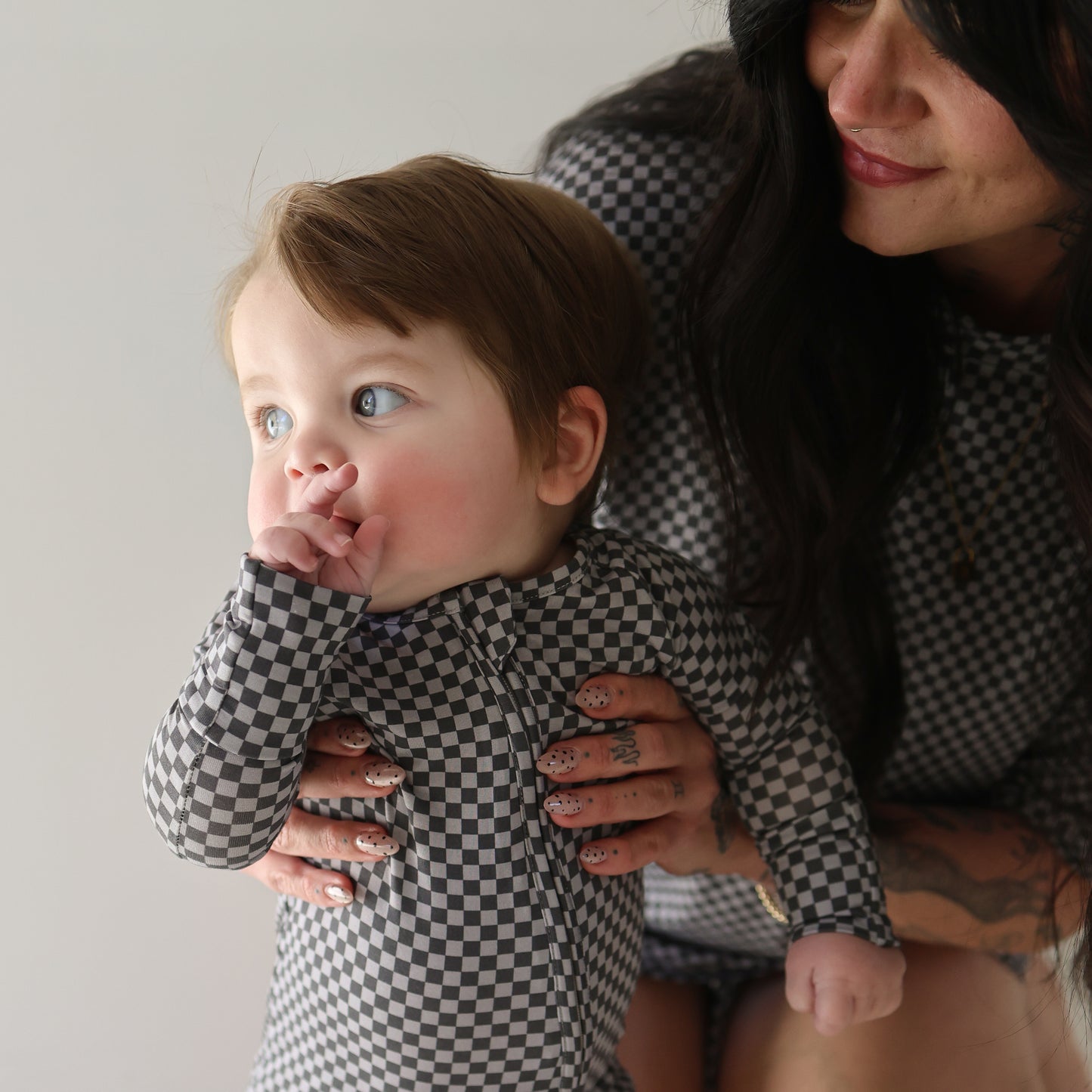 A baby wears forever french baby Bamboo Zip Pajamas in Street Micro Check and sucks their thumb while being held by a woman with long dark hair in matching checkered clothing and dotted nails. Both gaze off to the side.