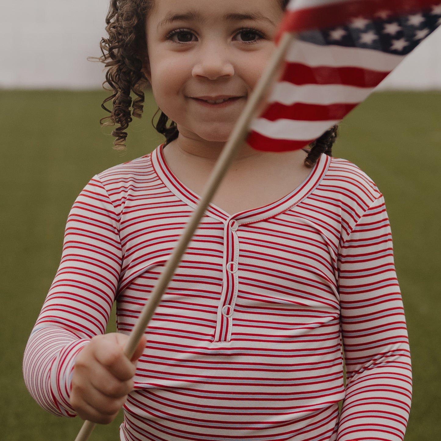 A young child with curly hair smiles outdoors on a grassy field, wearing forever french baby's Bamboo Two Piece Pajamas in the Heritage Stripe pattern and holding an American flag.