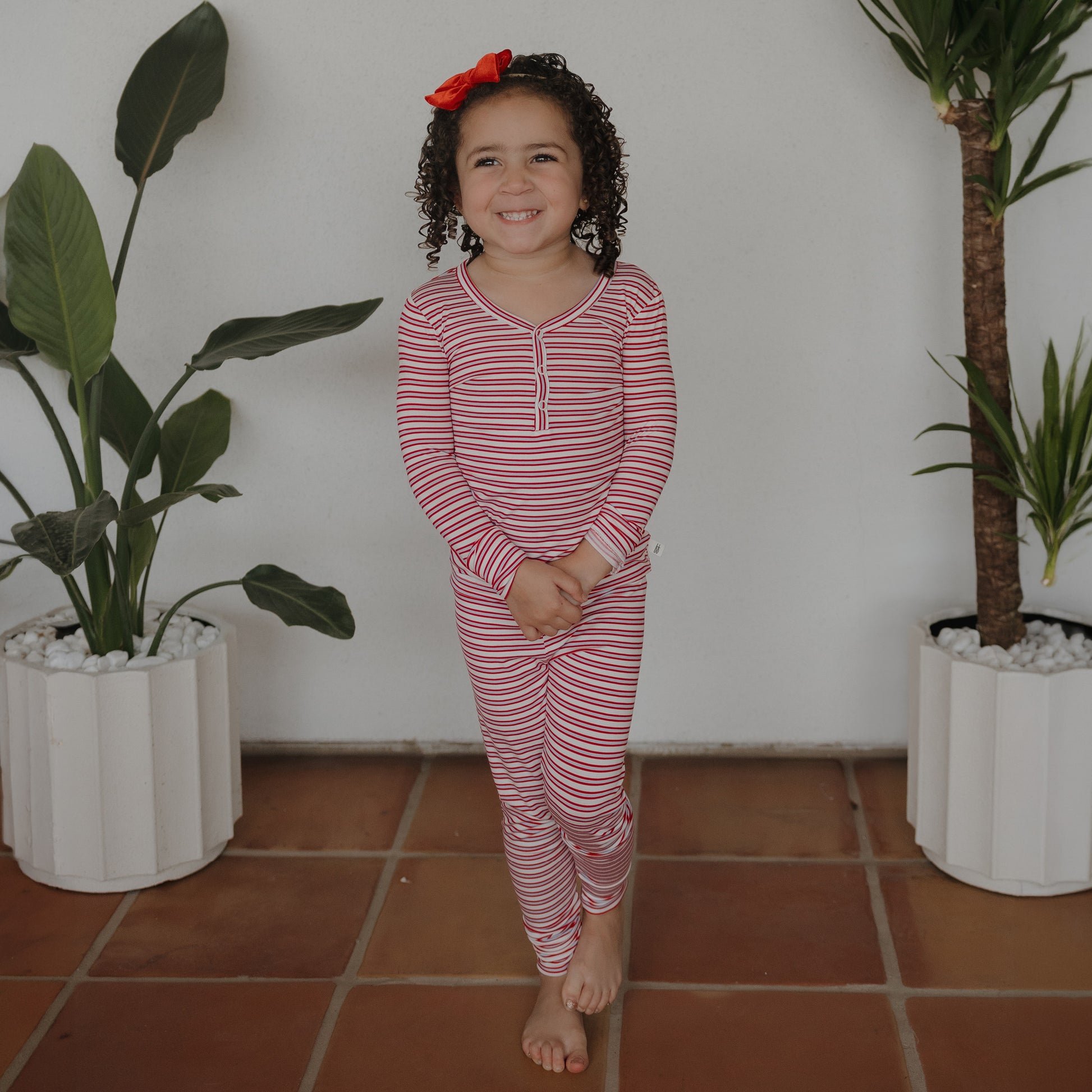 A young girl with curly hair and a red bow smiles at the camera, standing barefoot between two potted plants and wearing forever french baby’s Bamboo Two Piece Pajamas in Heritage Stripe.