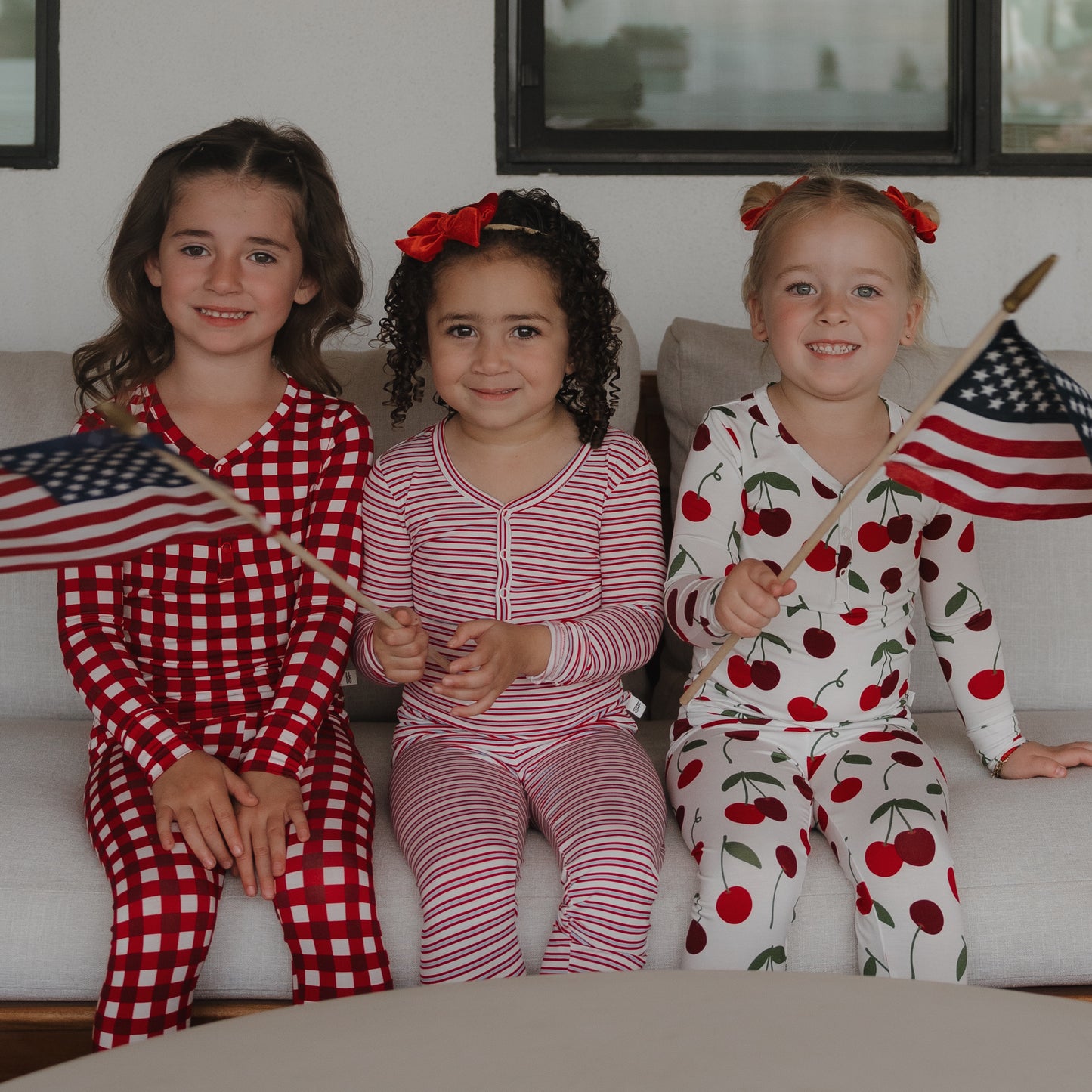 Three young girls in hypo-allergenic forever french baby Bamboo Two Piece Pajamas | Heritage Stripe sit on a couch, smiling. Two hold small American flags, and their festive sleepwear creates a playful, patriotic vibe.