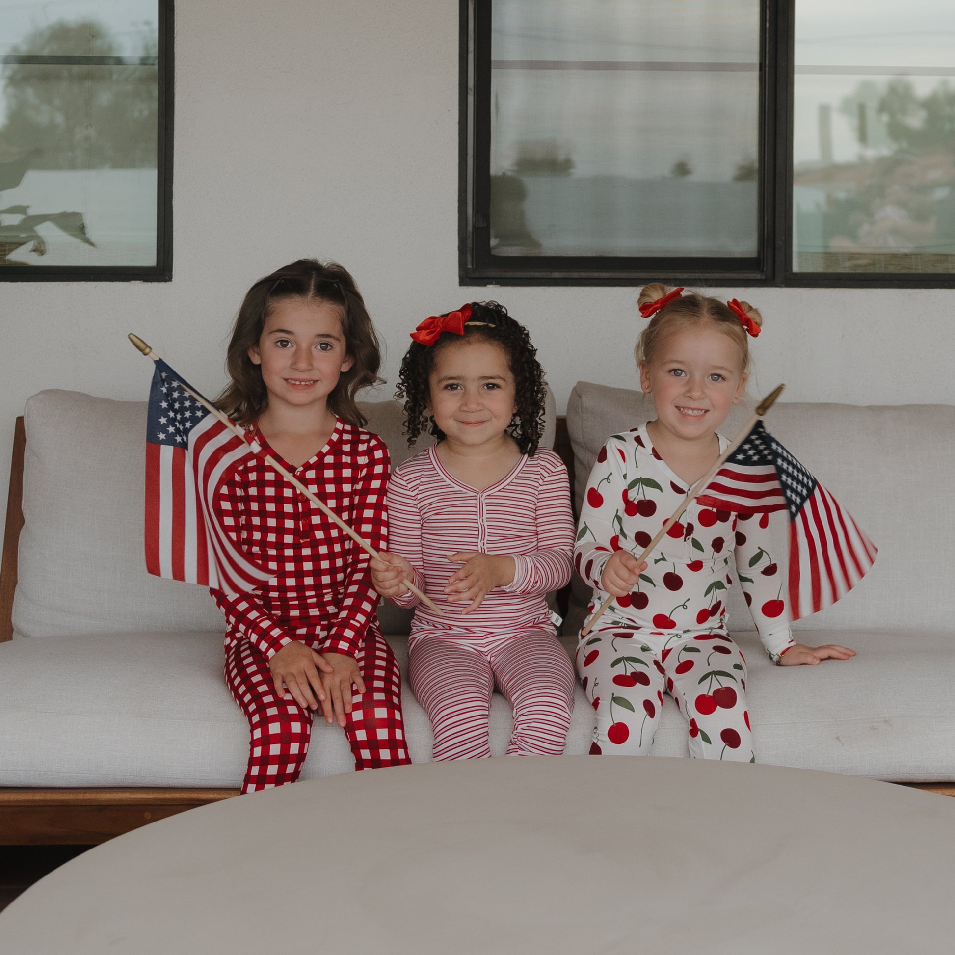 Three young girls smile on a couch holding American flags, dressed in forever french baby Bamboo Two Piece Pajamas | Heritage Stripe and festive outfits, with red bows in their hair. A table sits in front of them.