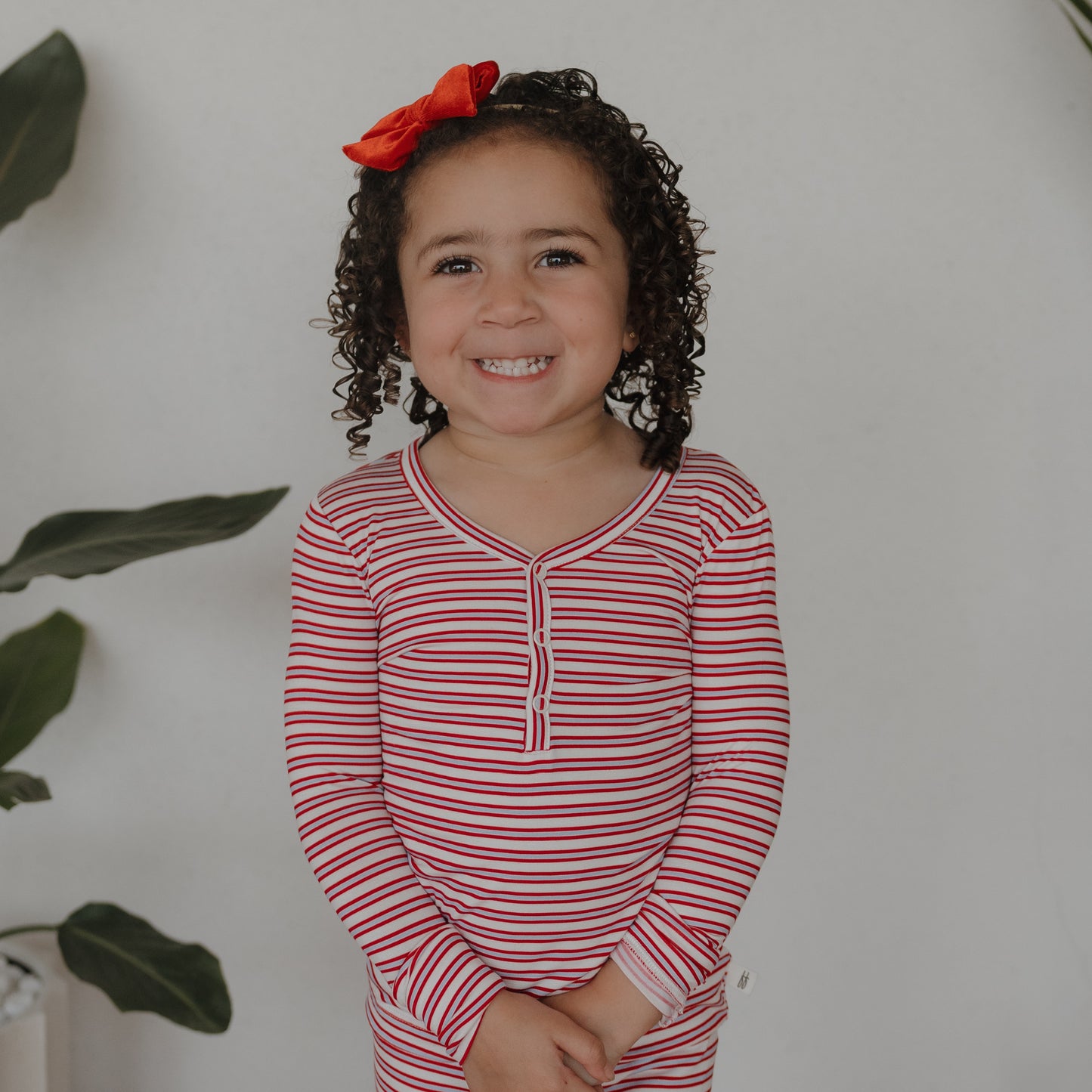 A young child with curly hair and a red bow smiles indoors against a white wall, wearing forever french baby’s Bamboo Two Piece Pajamas in Heritage Stripe. Green leafy plants are partially visible in the background.