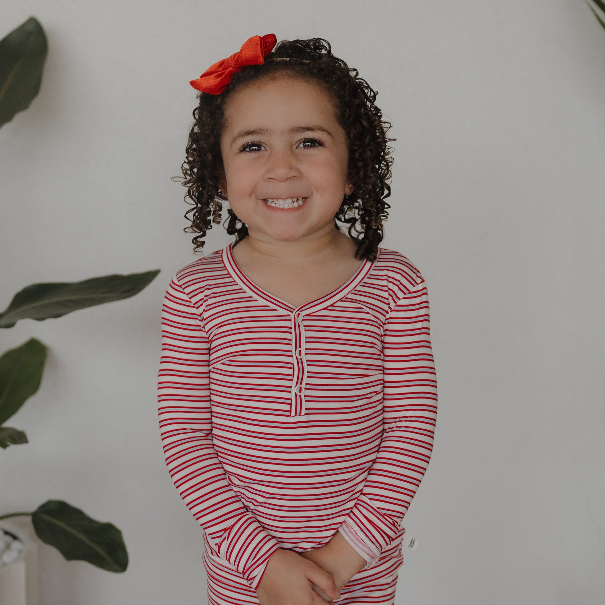 A young child with curly hair and a red bow smiles indoors against a white wall, wearing forever french baby’s Bamboo Two Piece Pajamas in Heritage Stripe. Green leafy plants are partially visible in the background.