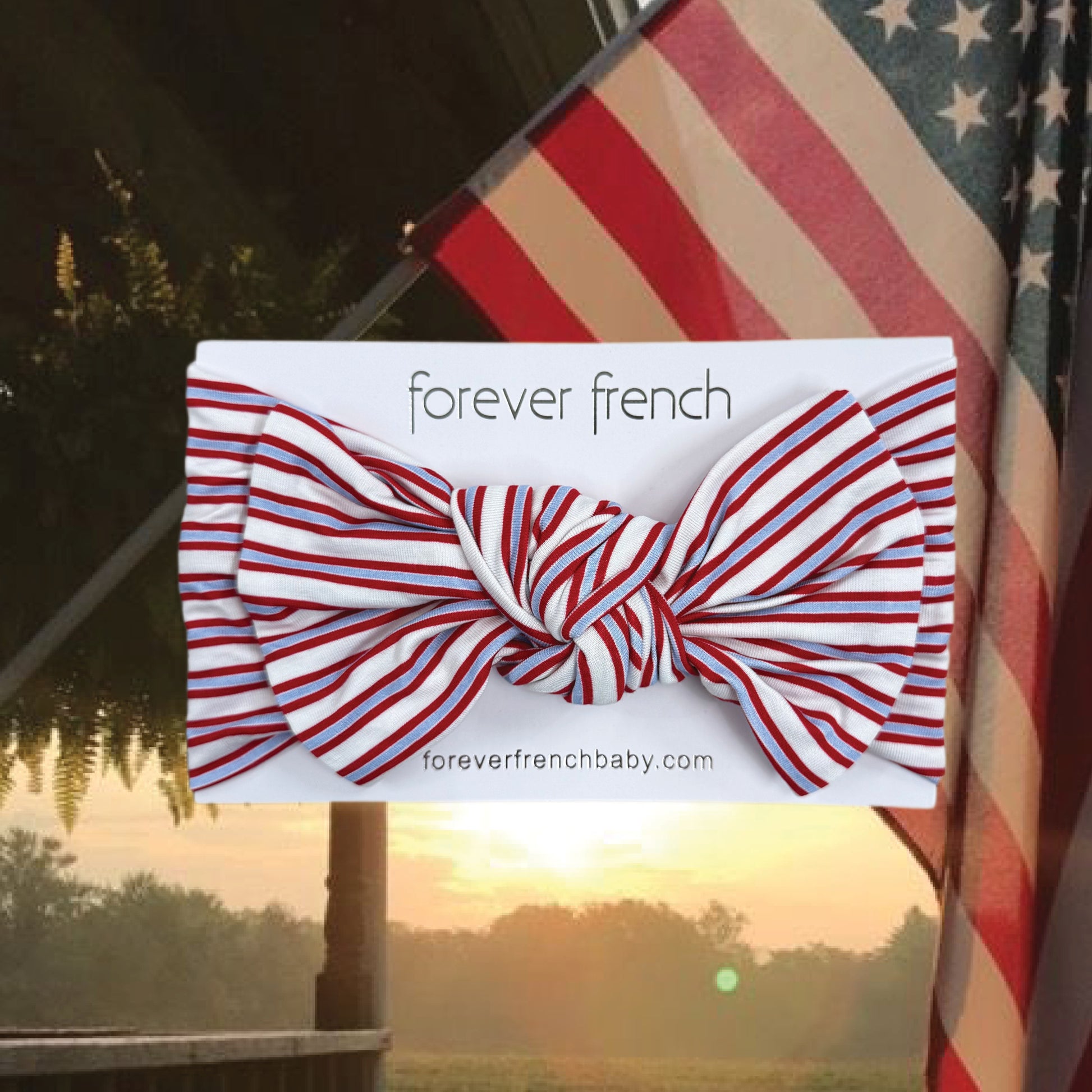 The Bamboo Head Wrap | Heritage Stripe from forever french baby, featuring red and white stripes, is displayed in front of a waving American flag at sunrise or sunset.