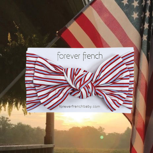 The Bamboo Head Wrap | Heritage Stripe from forever french baby, featuring red and white stripes, is displayed in front of a waving American flag at sunrise or sunset.