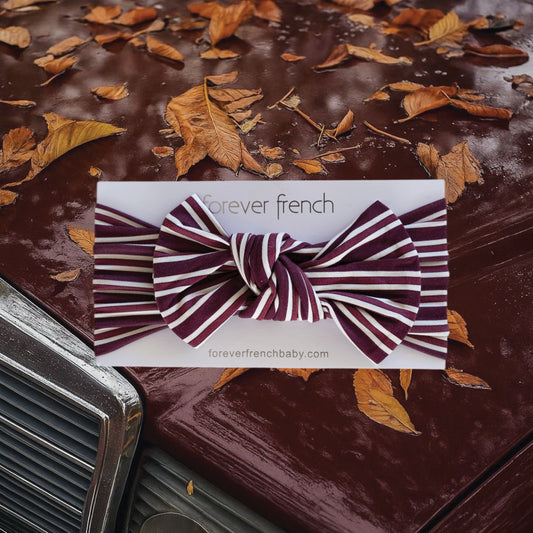 A bamboo head wrap in elegant maroon stripes from forever french baby is centered over a close-up of a vintage car hood adorned with raindrops and autumn leaves, evoking the cozy charm of fall. The Bamboo Head Wrap | Maroon Stripe adds a timeless touch to any little one’s ensemble.