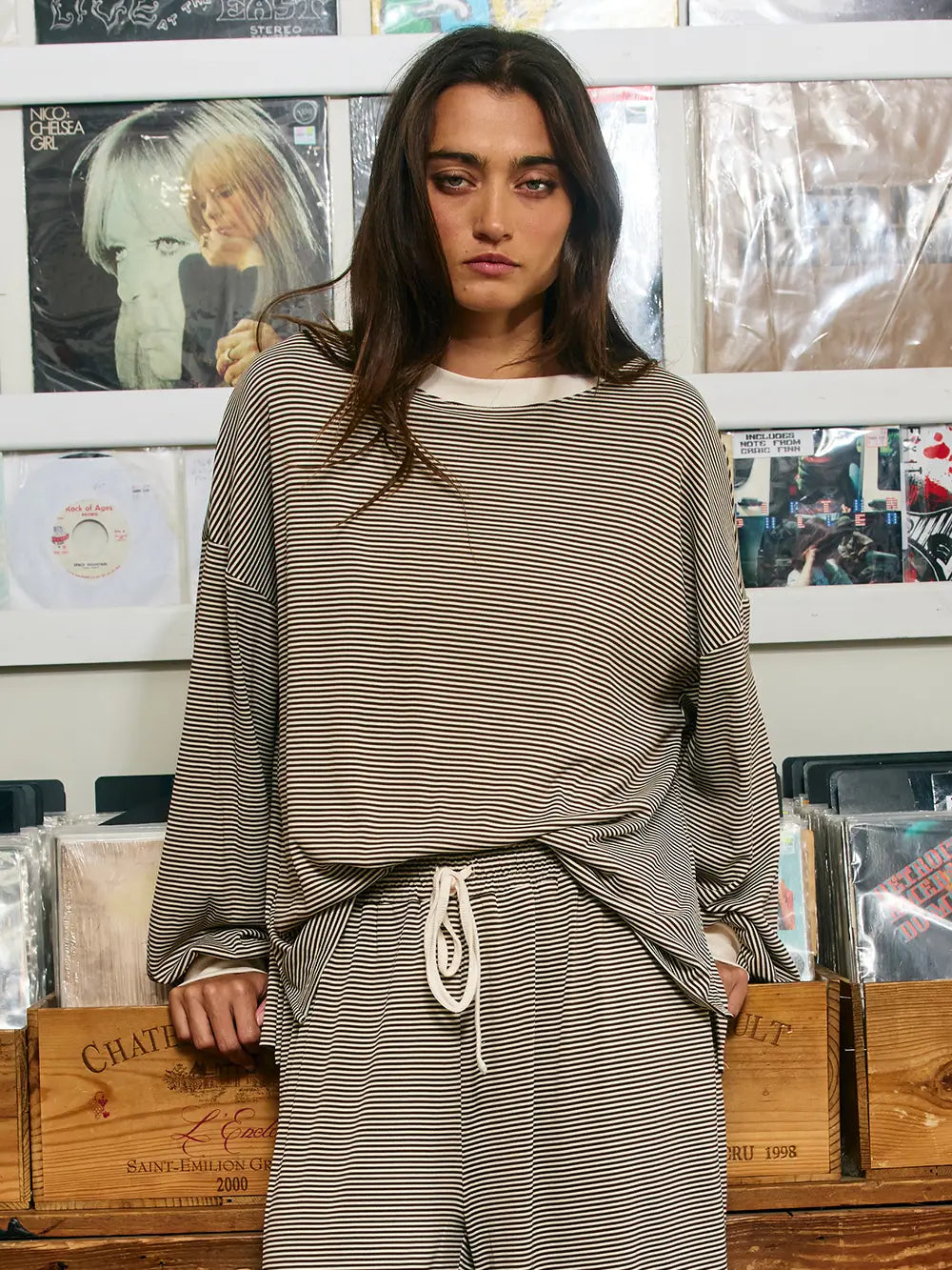 A woman wearing the bucket list Oversized Micro-Stripe Crewneck Top stands in front of a wall of vinyl records, looking at the camera with a neutral expression. Wooden crates filled with records are visible below.