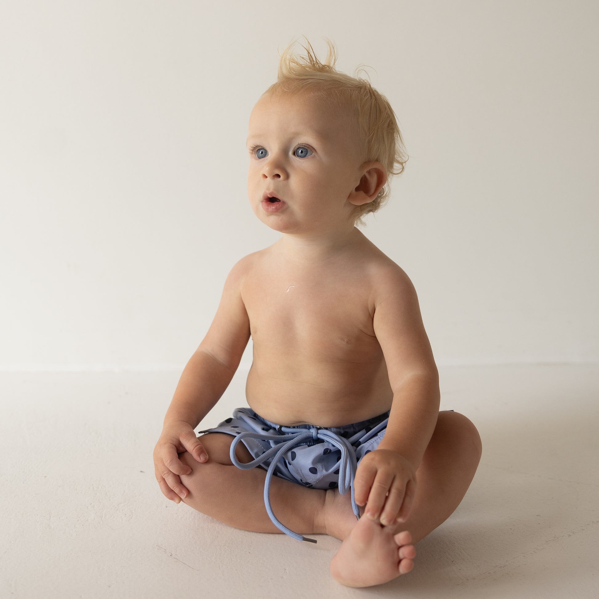 A blonde toddler with blue eyes sits cross-legged on a neutral background, wearing forever french baby’s Child Board Short | Sully and looking slightly upward with an open-mouthed expression.