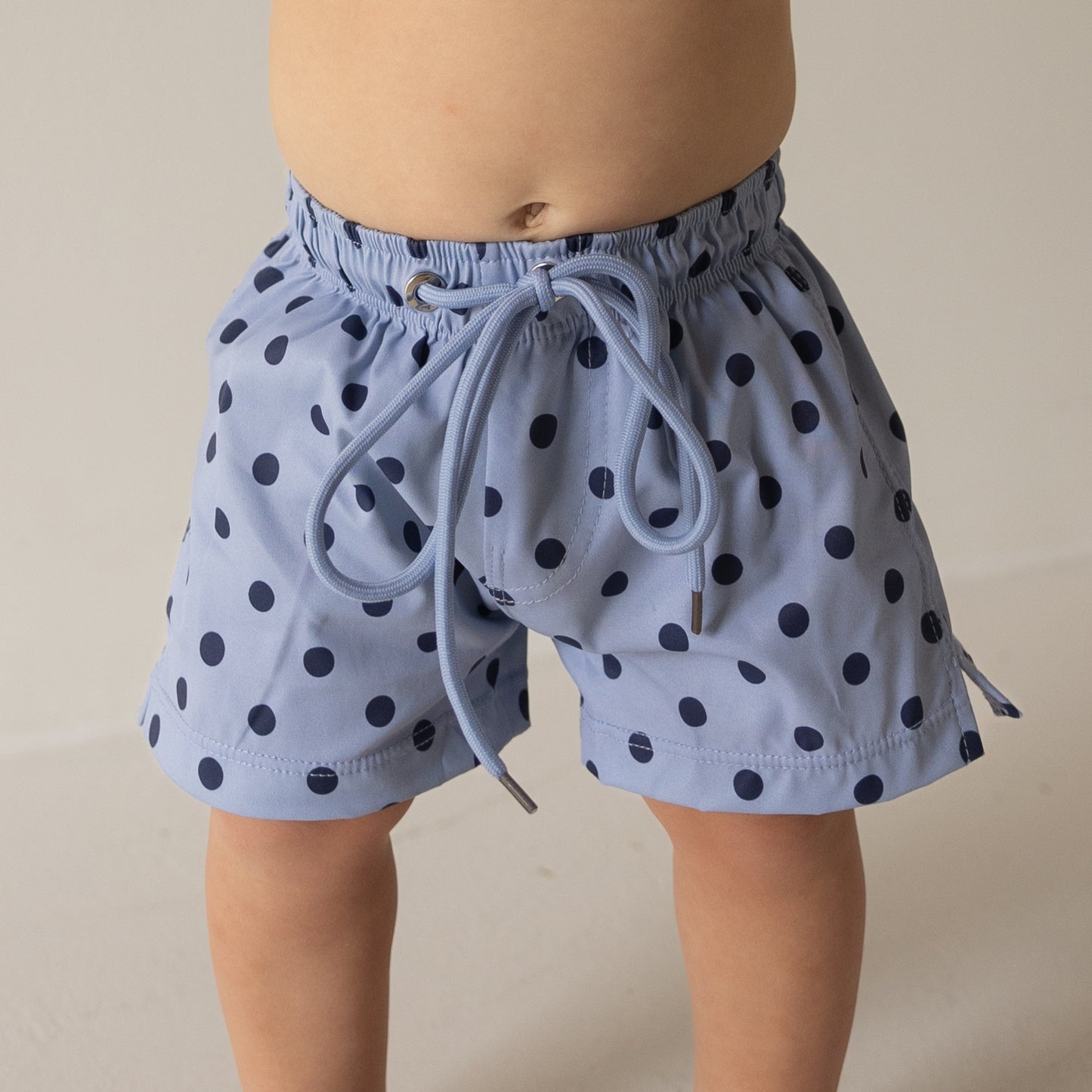 A young child stands against a plain light background wearing the forever french baby Child Board Short | Sully—light blue kids' swimwear shorts with dark blue polka dots and a drawstring waistband.