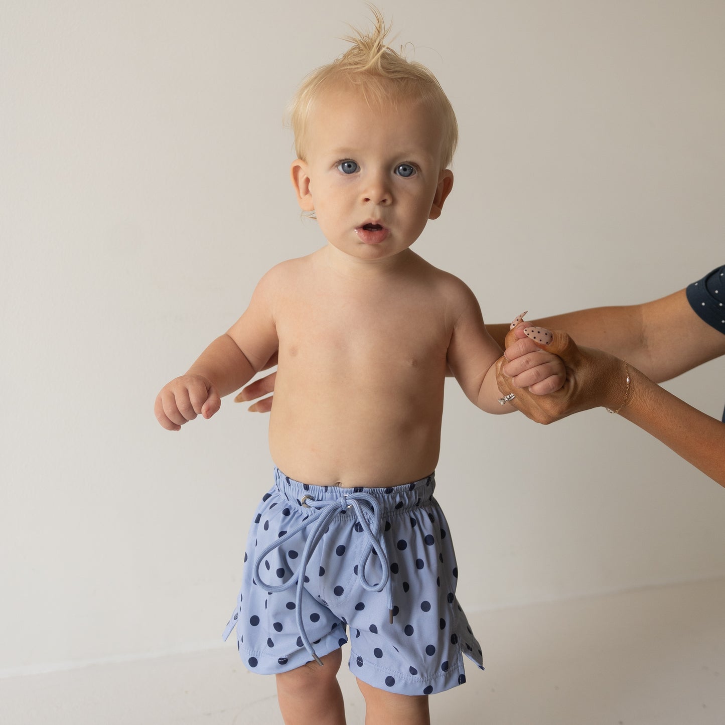 A toddler in forever french baby's Sully Child Board Short stands against a light background, holding an adult's hand—ideal for highlighting matching family looks or cute kids' swimwear.