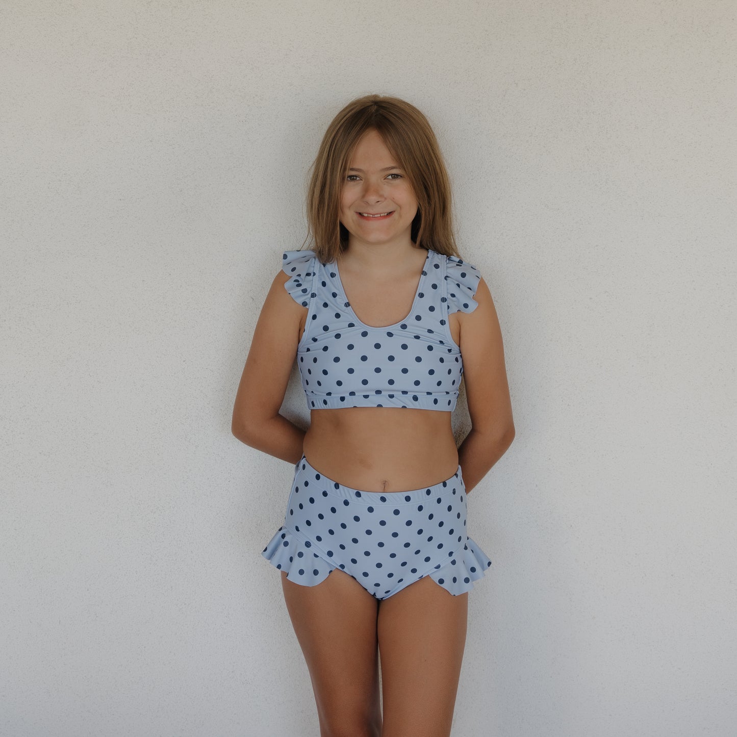 A smiling girl with light brown hair stands before a white wall, wearing the forever french baby Girl's Two Piece Swimsuit | Sully, featuring blue polka dots and playful ruffled details on the shoulders and hips.