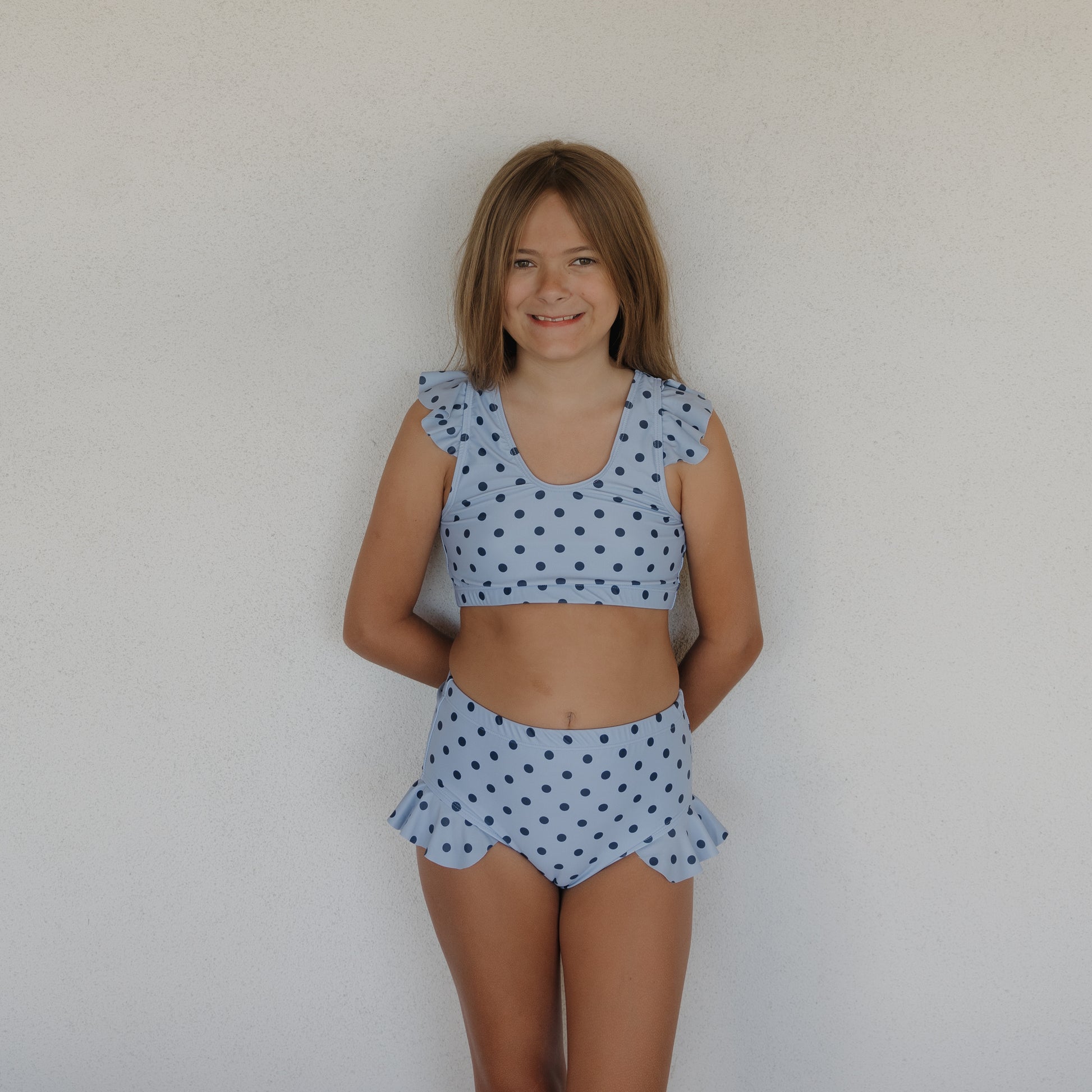 A smiling girl with light brown hair stands before a white wall, wearing the forever french baby Girl's Two Piece Swimsuit | Sully, featuring blue polka dots and playful ruffled details on the shoulders and hips.