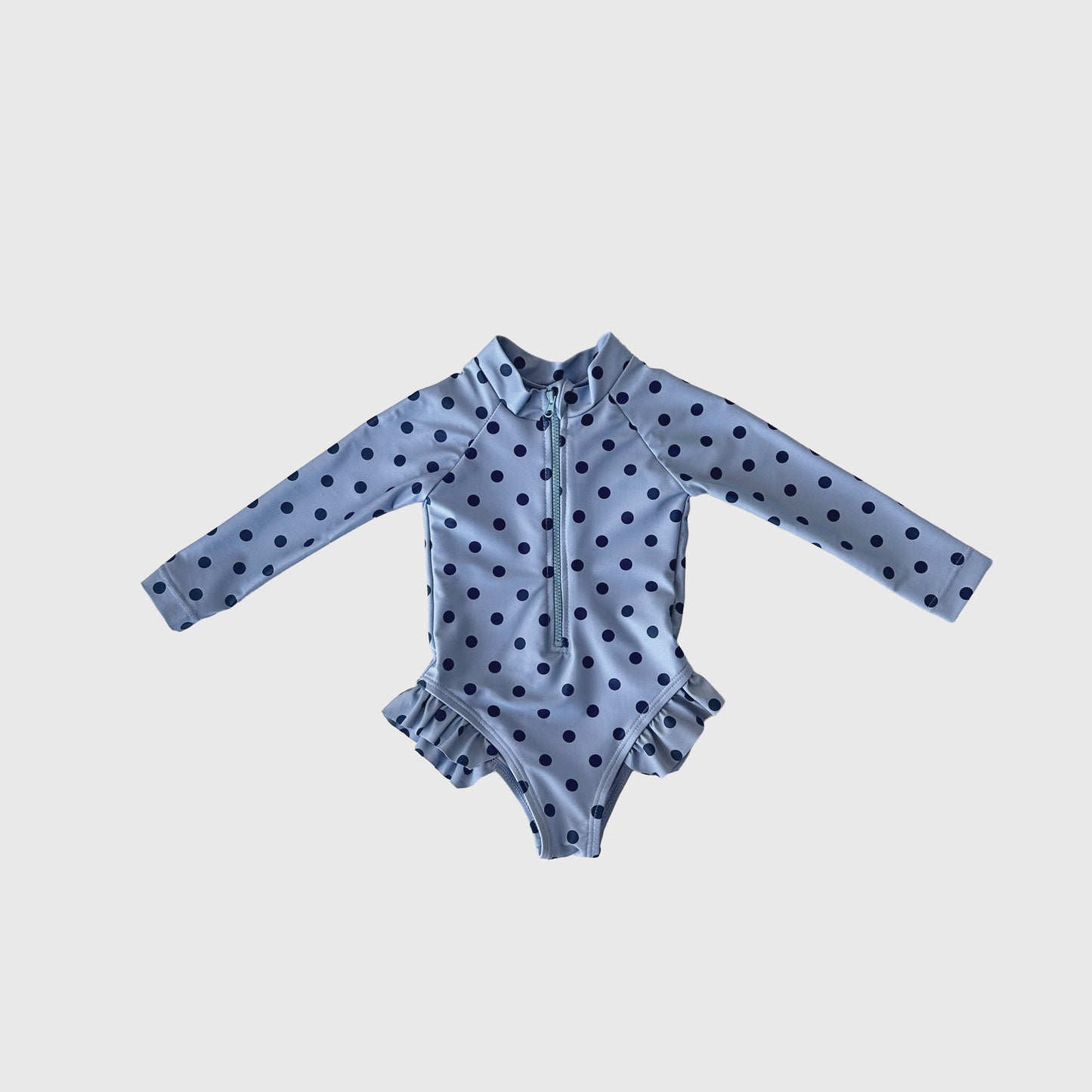 The Girl's Long Sleeve Swimsuit | Sully by forever french baby features light blue fabric with dark blue polka dots, a front zipper, and hip ruffles—ideal for matching family swimwear—shown on a plain white background.