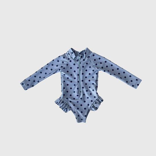 The Girl's Long Sleeve Swimsuit | Sully by forever french baby features light blue fabric with dark blue polka dots, a front zipper, and hip ruffles—ideal for matching family swimwear—shown on a plain white background.