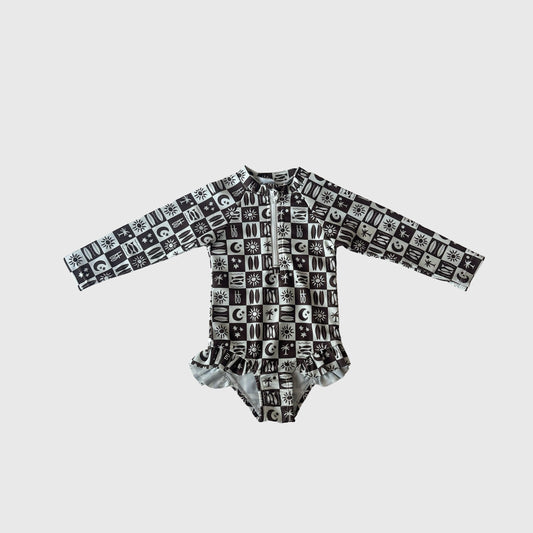 The Girl's Long Sleeve Swimsuit | Summer Check by forever french baby features a black and white grid pattern with sun, moon, and star symbols plus a ruffled waist—an adorable swim-ready outfit for kids.