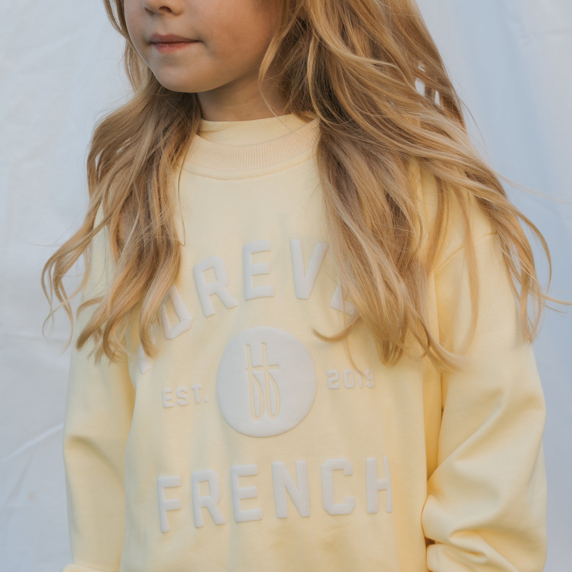 A young girl with long blonde hair wears the Child Sweatshirt | ff Yellow by forever french baby, made from 100% cotton and featuring "FOREVER FRENCH EST. 2019" text, standing against a light background and looking slightly to the side.
