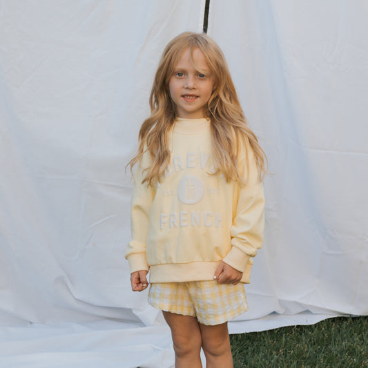 A young girl with long blonde hair stands on grass before a white backdrop, wearing the ff Yellow Child Sweatshirt by forever french baby and yellow plaid shorts. She smiles softly at the camera—ideal for matching family outfits.