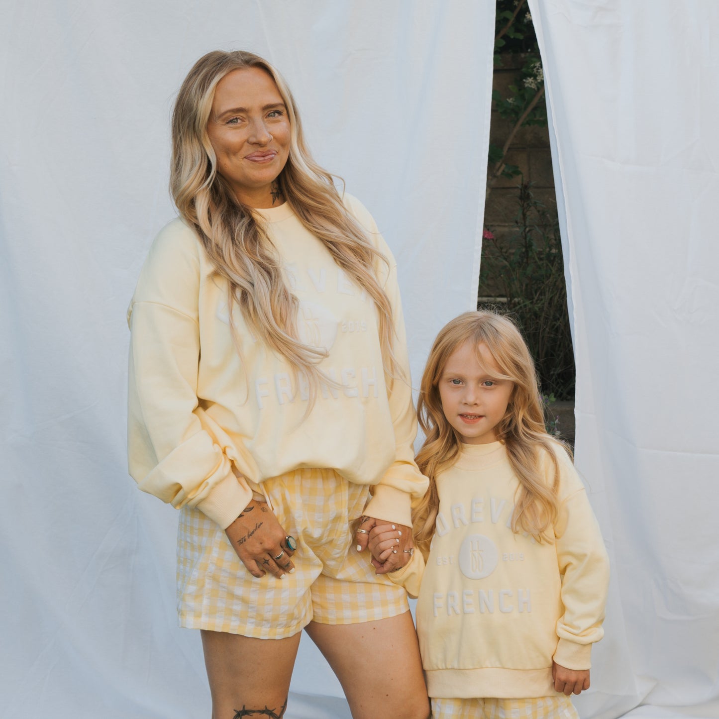 A woman and a young girl hold hands outdoors, both wearing matching "Child Sweatshirt | ff Yellow" by forever french baby with checkered shorts, posing in front of a white cloth backdrop.