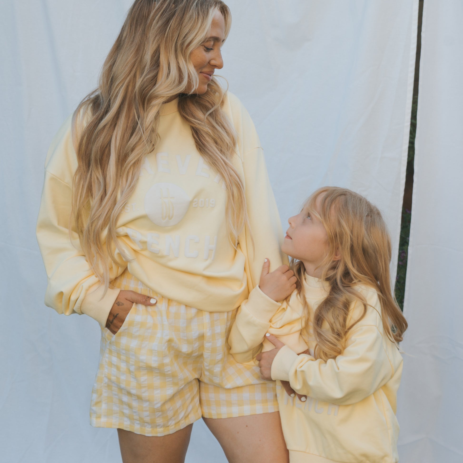 A woman and a young girl with long blonde hair smile at each other in matching pastel yellow outfits. The child wears the forever french baby "Child Sweatshirt | ff Yellow," made from 100% cotton, against a white background.