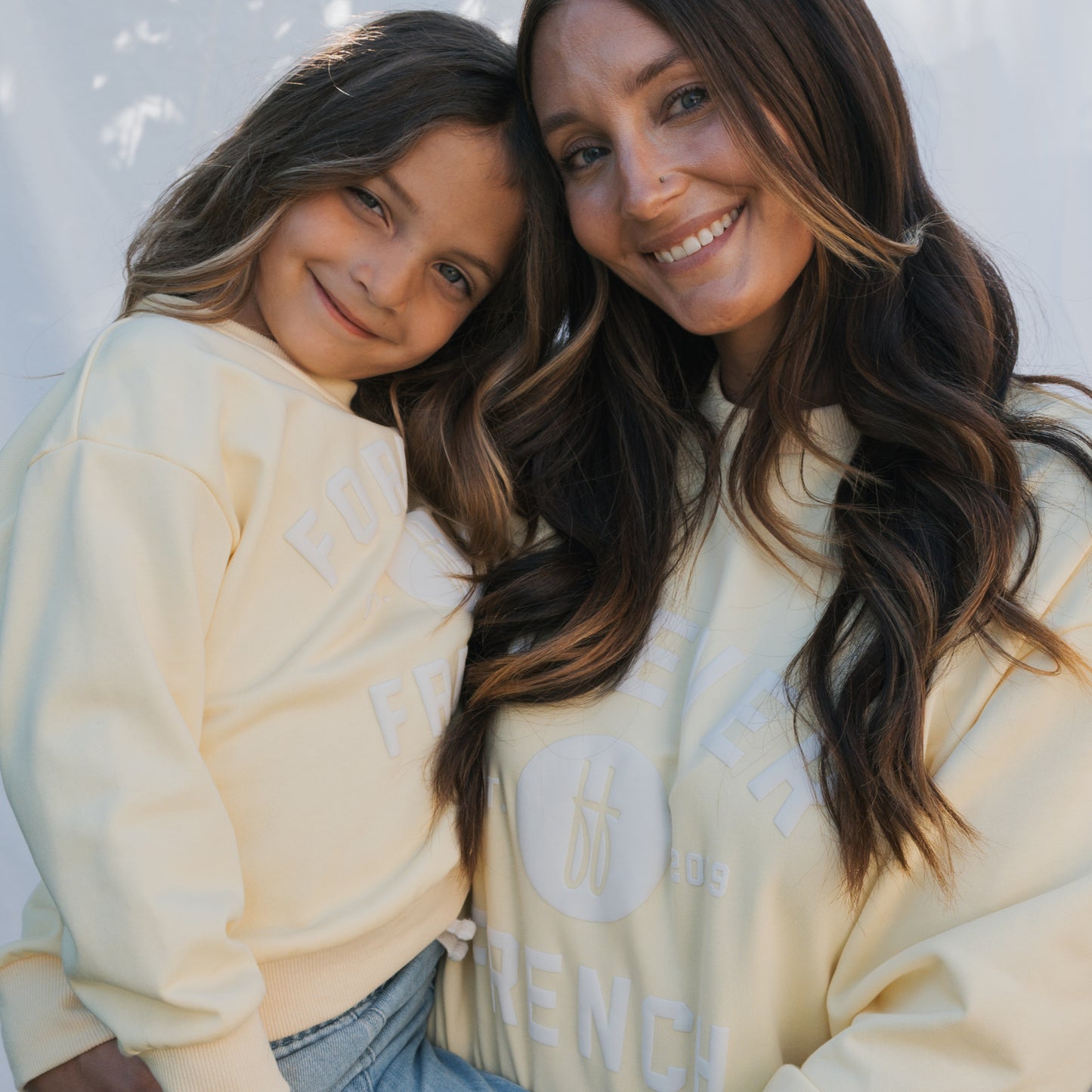 A woman and a young girl smile at the camera, both wearing matching "Child Sweatshirt | ff Yellow" by forever french baby—100% cotton and perfect for coordinated family outfits against a soft, bright background.