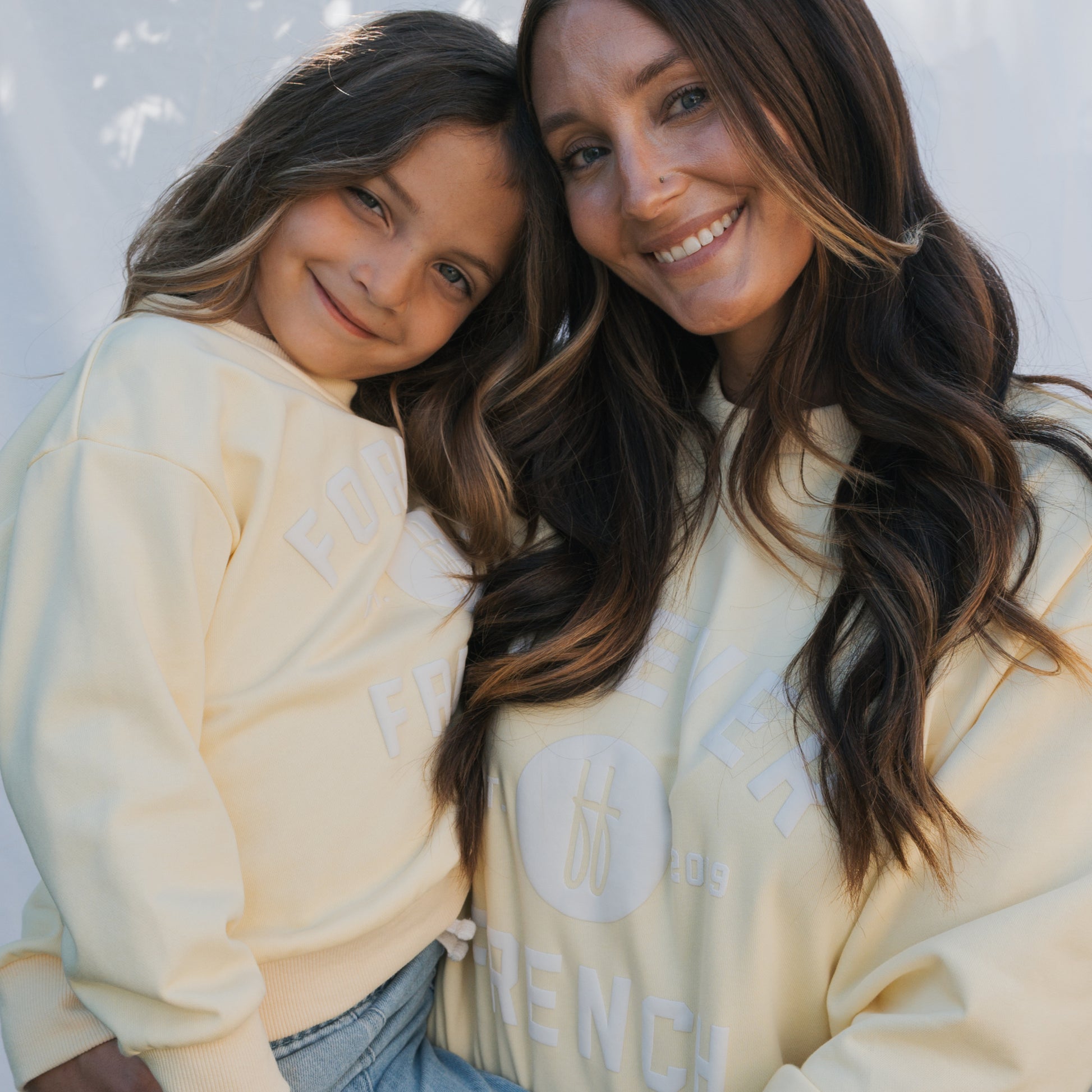 A woman and a young girl smile at the camera, both wearing matching "Child Sweatshirt | ff Yellow" by forever french baby—100% cotton and perfect for coordinated family outfits against a soft, bright background.