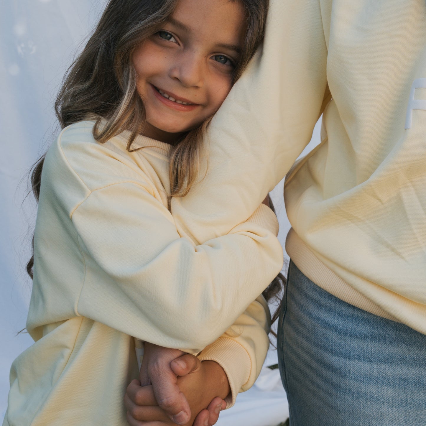 A young girl with long brown hair smiles while hugging an adult, both looking happy and close. She wears the forever french baby Child Sweatshirt | ff Yellow, paired with jeans, against a light background in matching family outfits.
