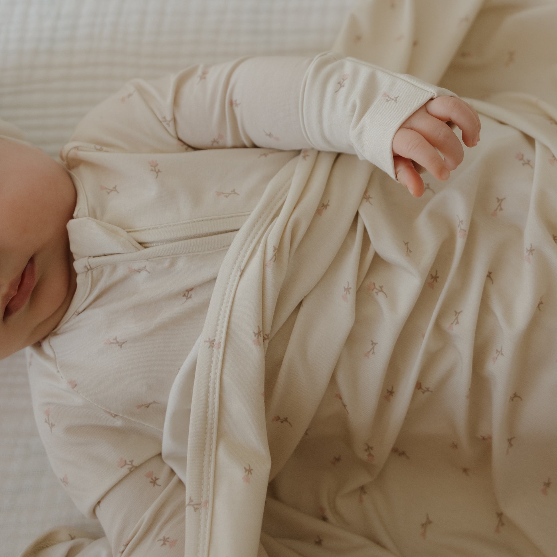 A baby in a soft, cream-colored onesie with star patterns lies on the forever french baby Bamboo Swaddle | Lucy. Only part of the baby's face and one arm are visible.