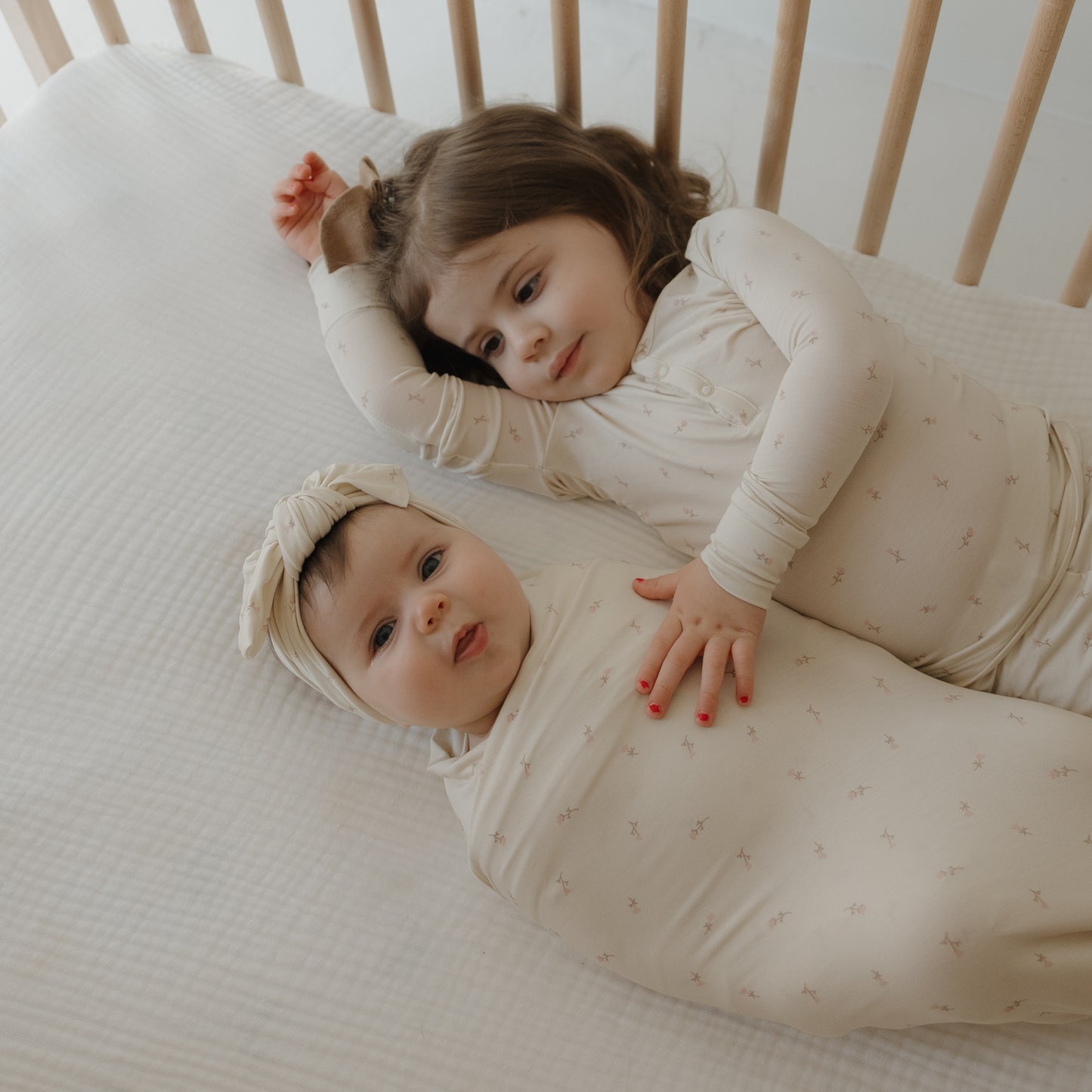 Two children rest in a crib: an older child in pajamas gently touches a baby wrapped in the forever french baby Bamboo Swaddle | Lucy with a matching headband, both lying on a soft, hypo-allergenic blanket.