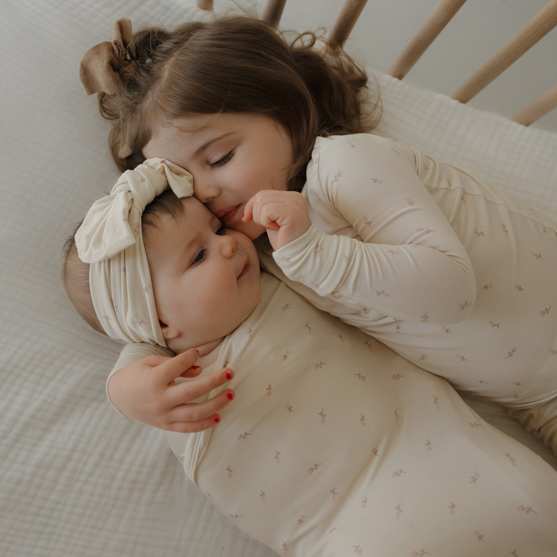 Two young children in matching cream pajamas cuddle on a bed. The older hugs and kisses the baby, who’s wrapped in the forever french baby Bamboo Swaddle | Lucy, looking up at her sibling. Both are cozy and comfortable.