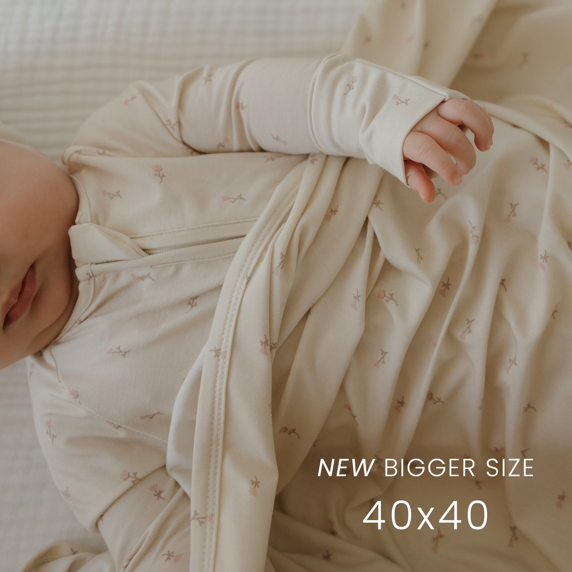 A baby in the forever french baby Bamboo Swaddle | Lucy, featuring light cream color and subtle patterns, rests on a white blanket. Text reads: "NEW BIGGER SIZE 40x40." This breathable wrap keeps your little one comfy. Baby’s face is partially visible.