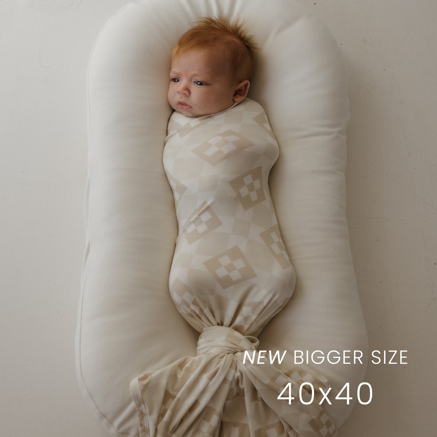 A red-haired baby lies swaddled in the forever french baby Bamboo Swaddle | Cottage Quilt on a beige cushioned lounger. Text reads "NEW BIGGER SIZE 40x40.