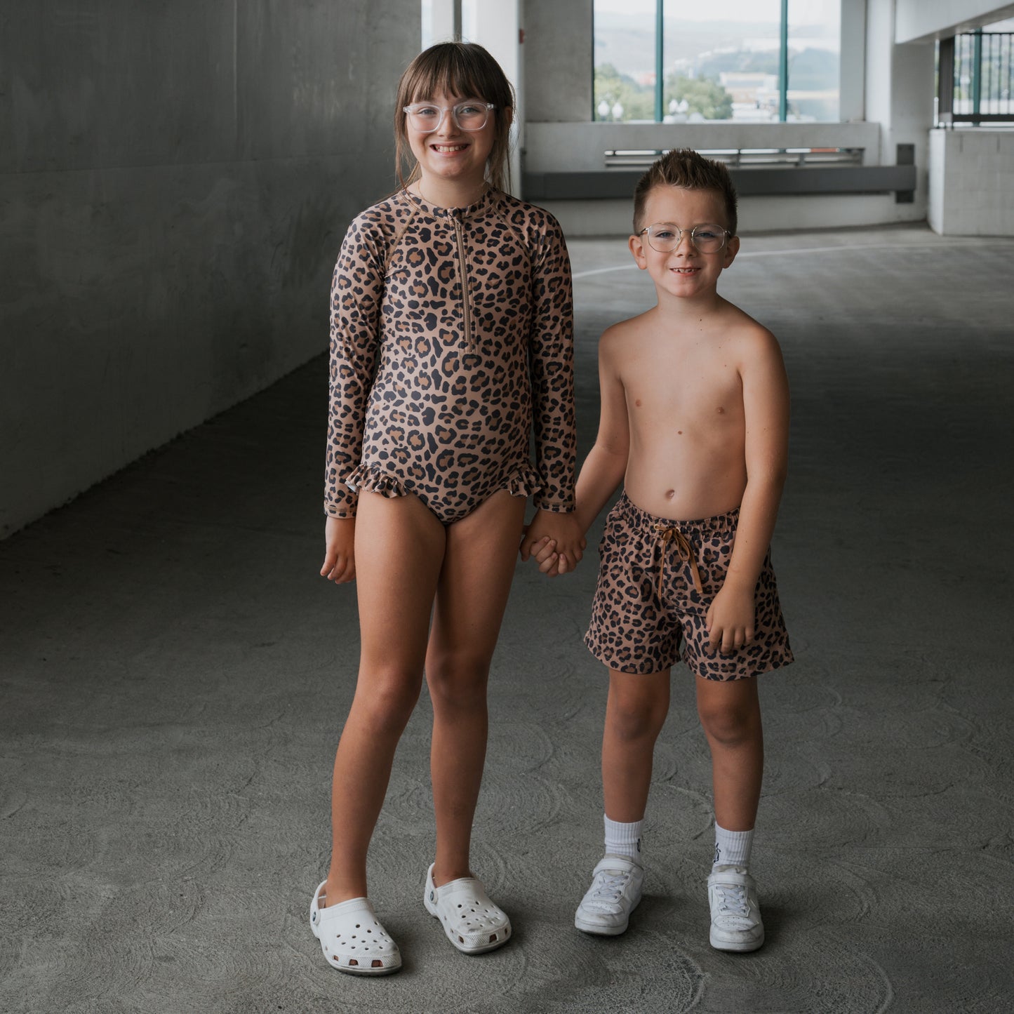 Two children wearing glasses pose indoors, smiling and holding hands. The girl wears a long-sleeve leopard print swimsuit and white clogs. The boy sports forever french baby *PRE-ORDER* Child Board Short | Leopard, white socks, and sandals.